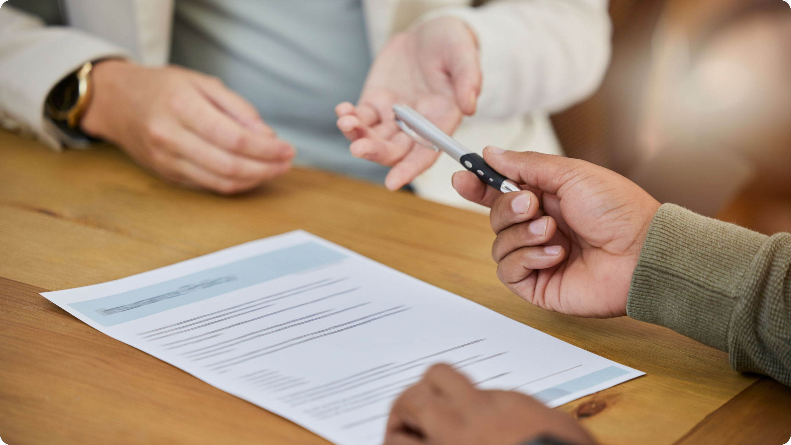A close-up of hands at a meeting table: one person holds a pen over a document, while another gestures. The document appears to be formal, possibly a contract or agreement, on a wooden table.