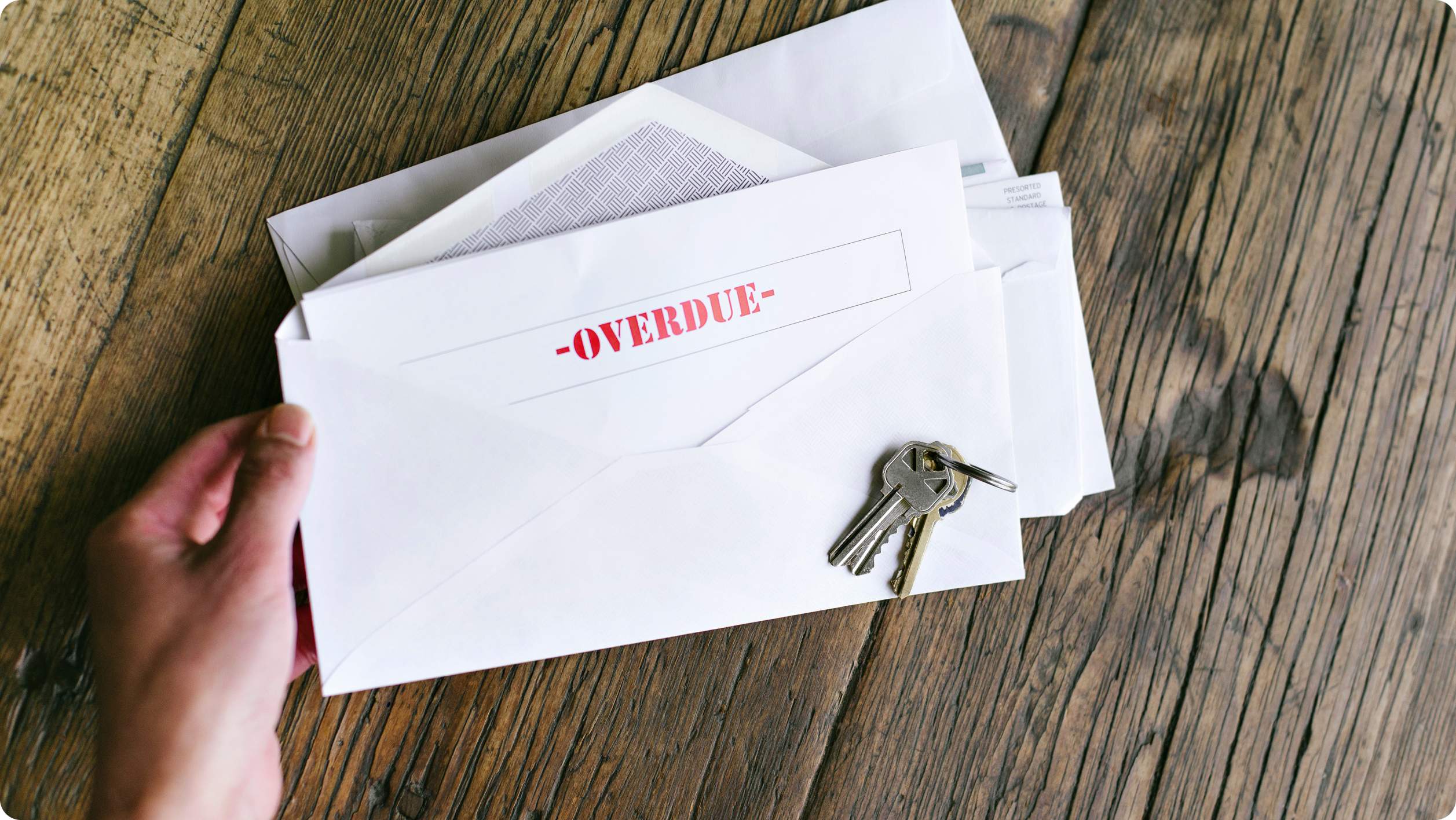 A hand holds several envelopes, with one prominently displaying the word "OVERDUE" in red. Two keys rest on top of the envelopes. The scene is set on a wooden surface.