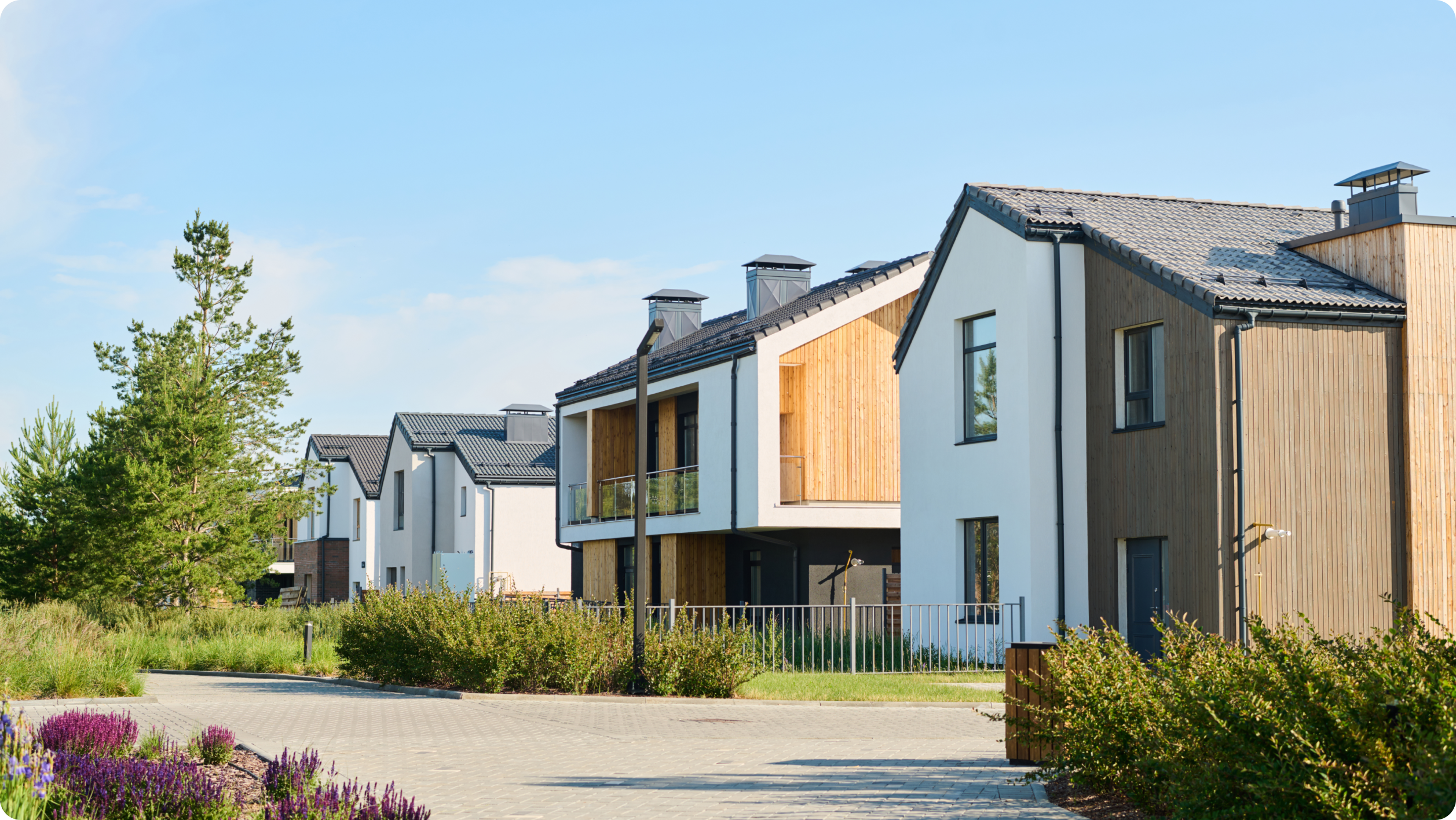A row of modern two-story houses with wooden accents and large windows. The homes have dark roofing and are surrounded by greenery and a paved pathway under a clear blue sky.