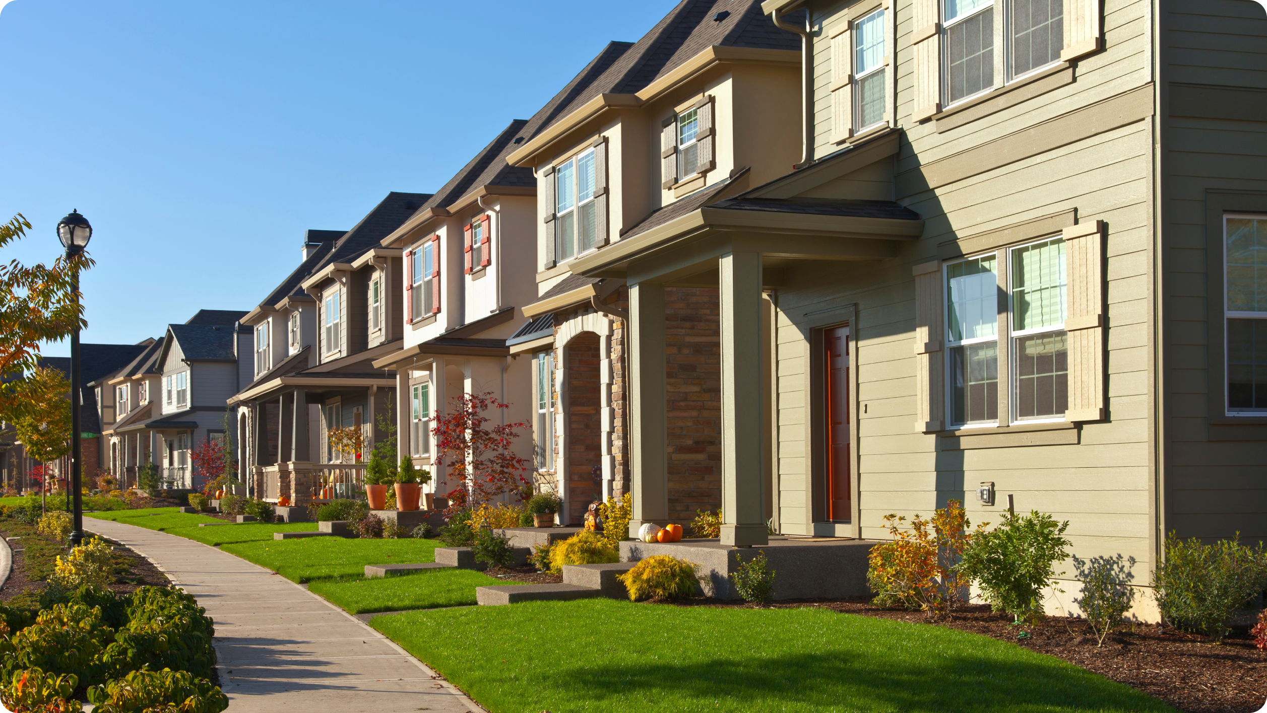 A row of modern suburban houses with well-maintained lawns and gardens on a sunny day. A sidewalk runs parallel to the homes, and the trees along the street have autumn foliage.