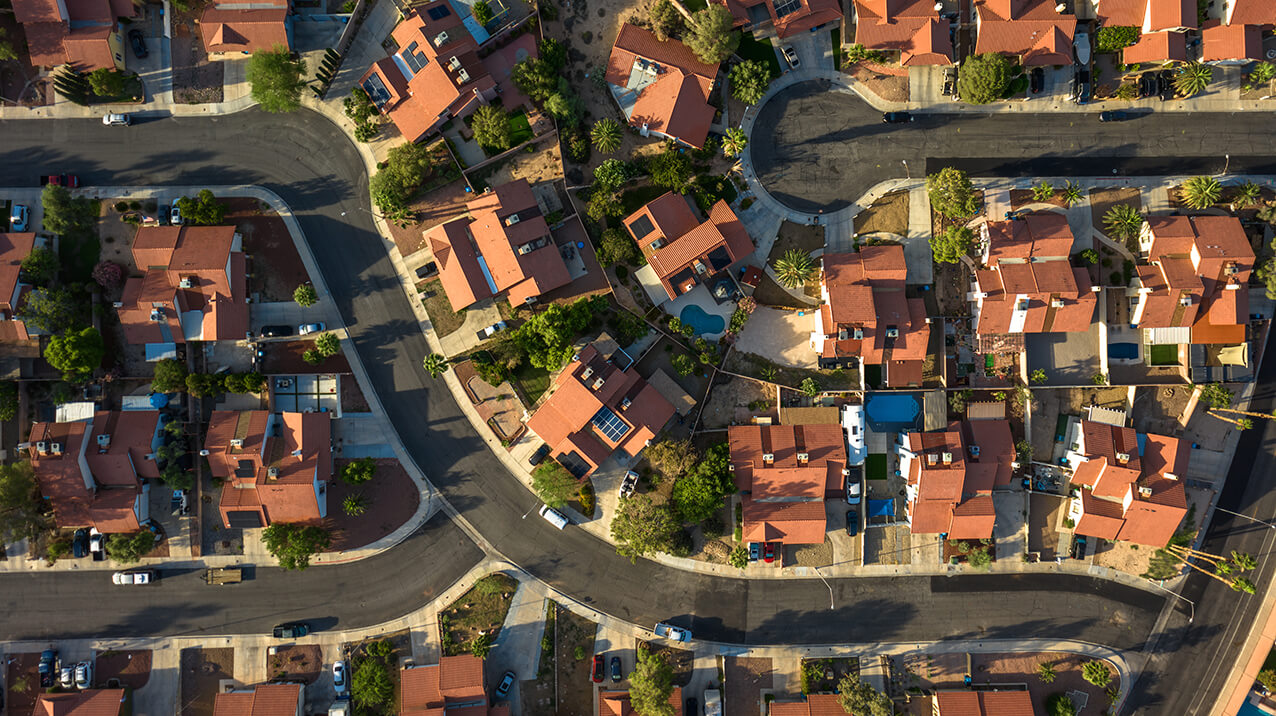 Aerial view of a suburban neighborhood with winding streets. The houses have red-tiled roofs and are surrounded by small yards and greenery. The layout shows a mix of curved and straight roads with parked cars visible along the streets.
