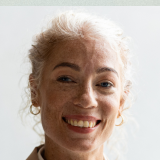 Smiling woman with light skin and curly white hair, wearing gold hoop earrings. She has a friendly expression and freckles, with a light background.
