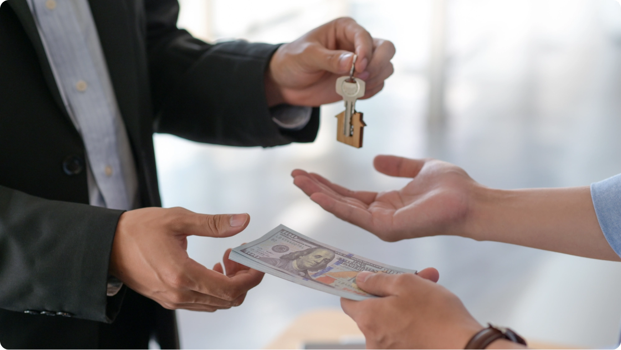 Two people are seen in a close-up exchange: one hands over a set of keys with a house-shaped keychain, while the other offers a stack of hundred-dollar bills. The background is blurred.