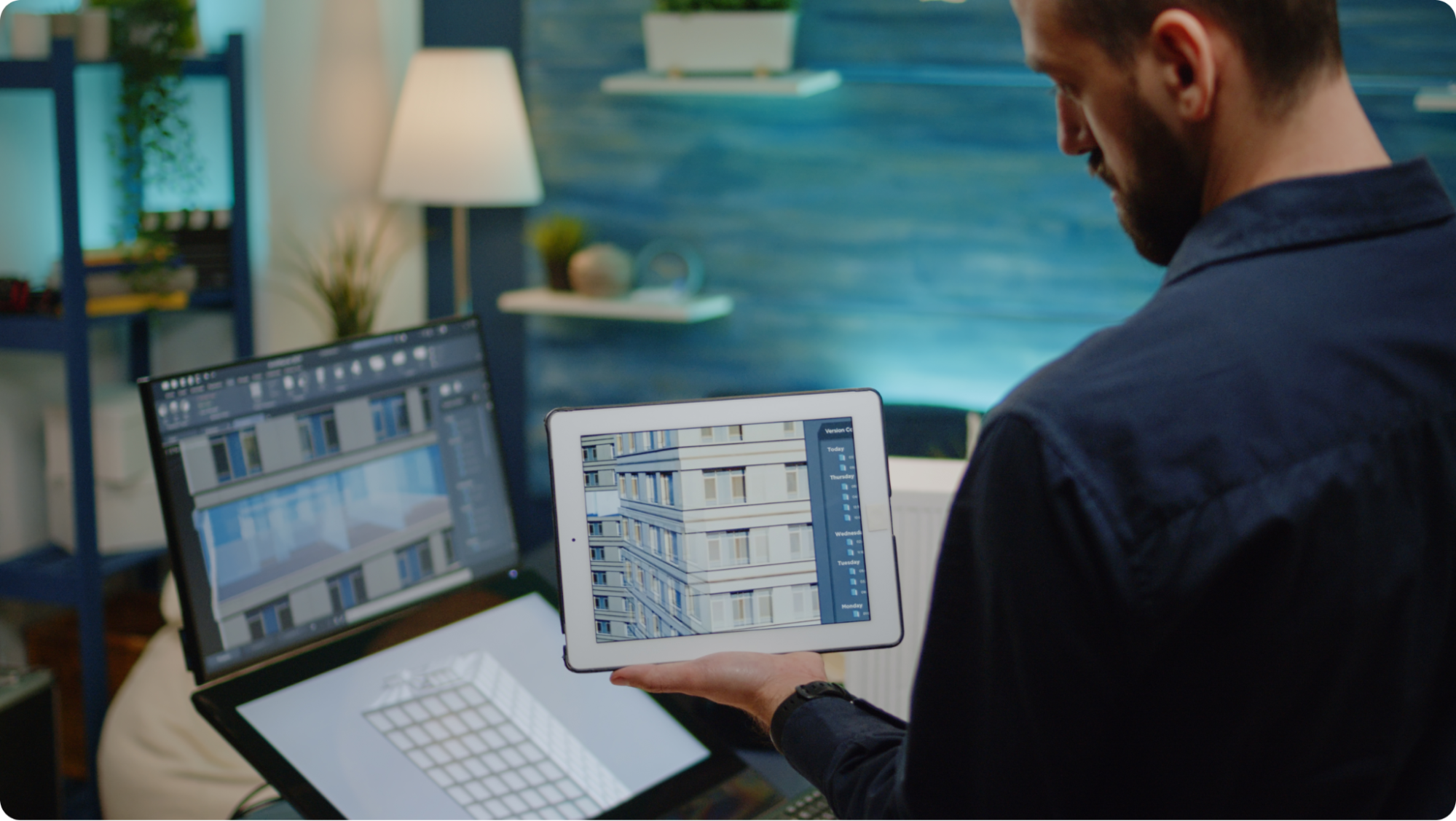 A person holding a tablet displaying architectural design software stands near two monitors showing detailed building designs. The workspace is lit warmly, with plants and a lamp in the background.