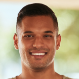 Smiling person with short dark hair, wearing a light-colored shirt, in an outdoor setting with blurred greenery in the background.