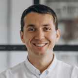 Smiling man with short dark hair wearing a white button-up shirt, standing indoors with a blurred background.