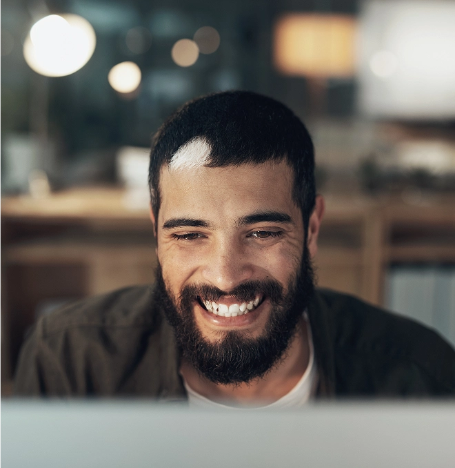 Man with a beard smiling while looking at a computer screen, seated in a dimly lit room with blurred background and warm lighting.