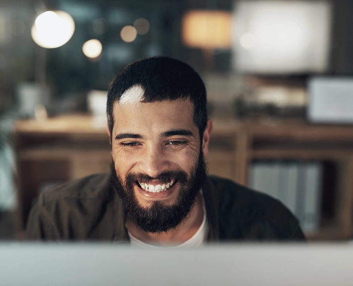A man with a beard is smiling while looking at a computer screen. The background is softly lit, suggesting a cozy, indoor setting with warm lights and blurred shelves.