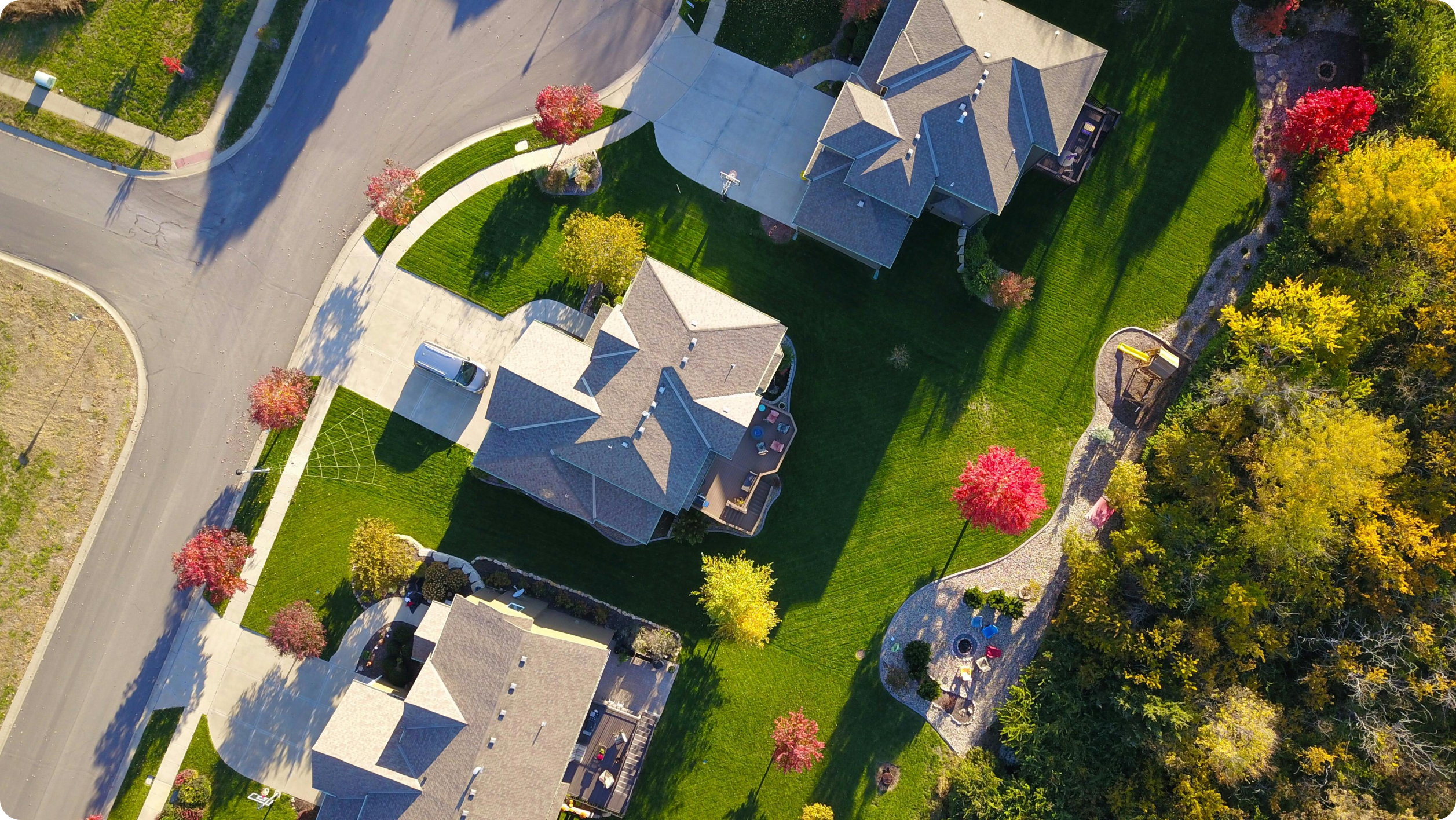 Aerial view of a suburban neighborhood with four houses. Each house has a driveway and well-manicured lawns. Vibrant autumn trees with red and yellow leaves add color. A curving road borders the front.