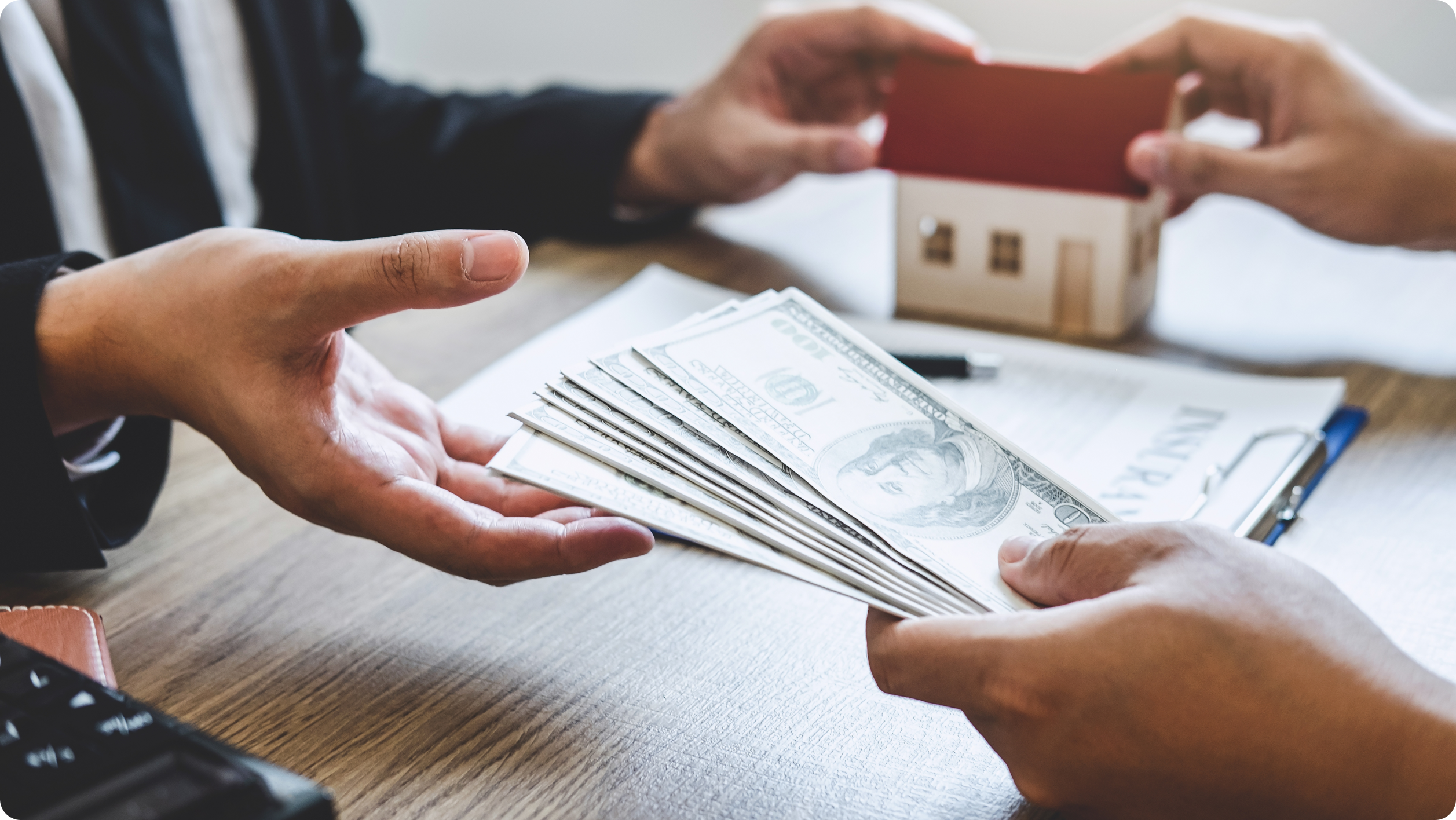 Two people sit at a wooden table exchanging U.S. dollar bills. One person holds a stack of money, while the other holds a small model house, seemingly discussing how to open an escrow account. A document with charts is visible on the table.