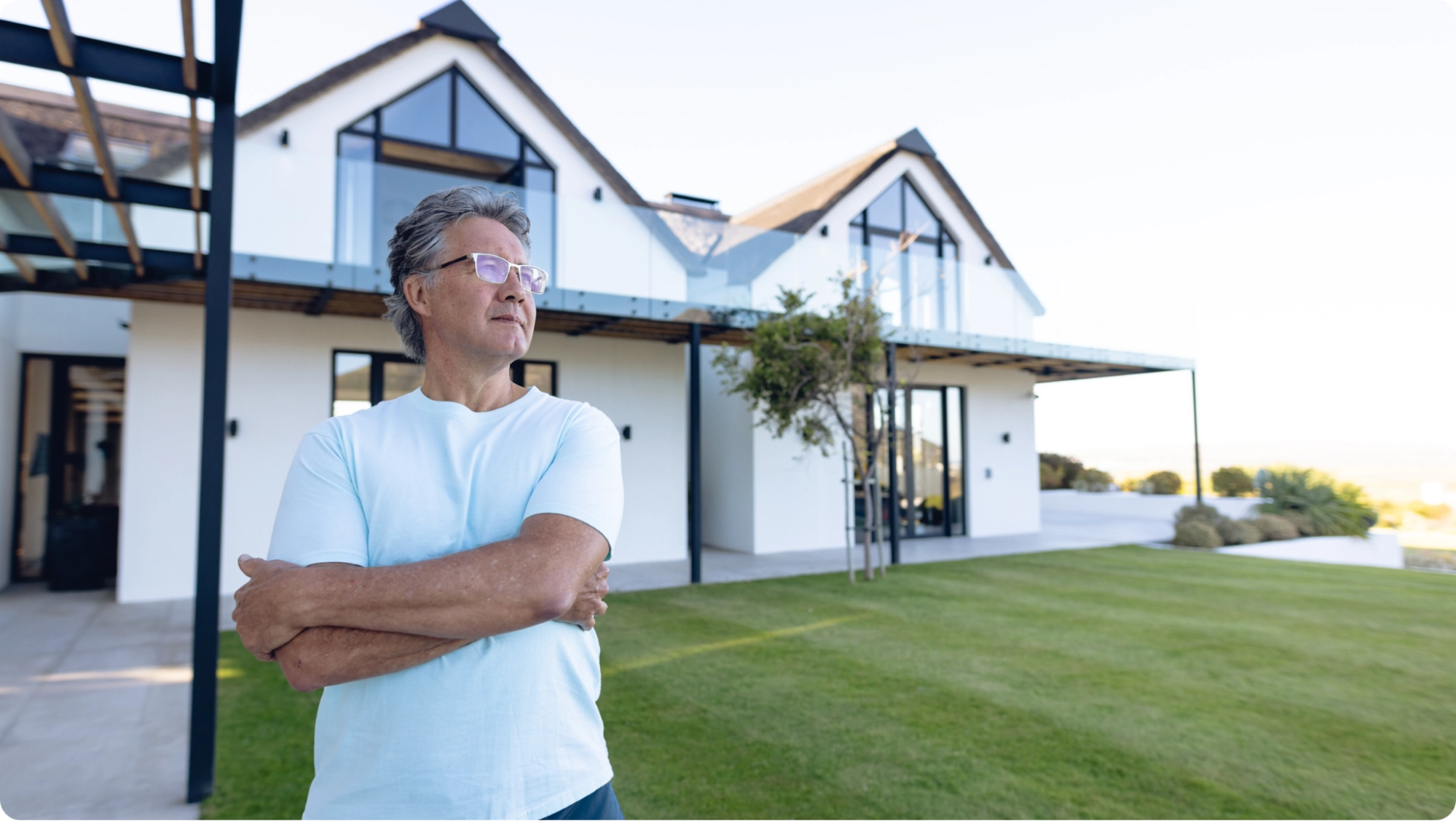 A person with gray hair and glasses stands outside, arms crossed, looking to the side. They are in front of a modern white house with a sloped roof and large windows, surrounded by a neatly manicured lawn.