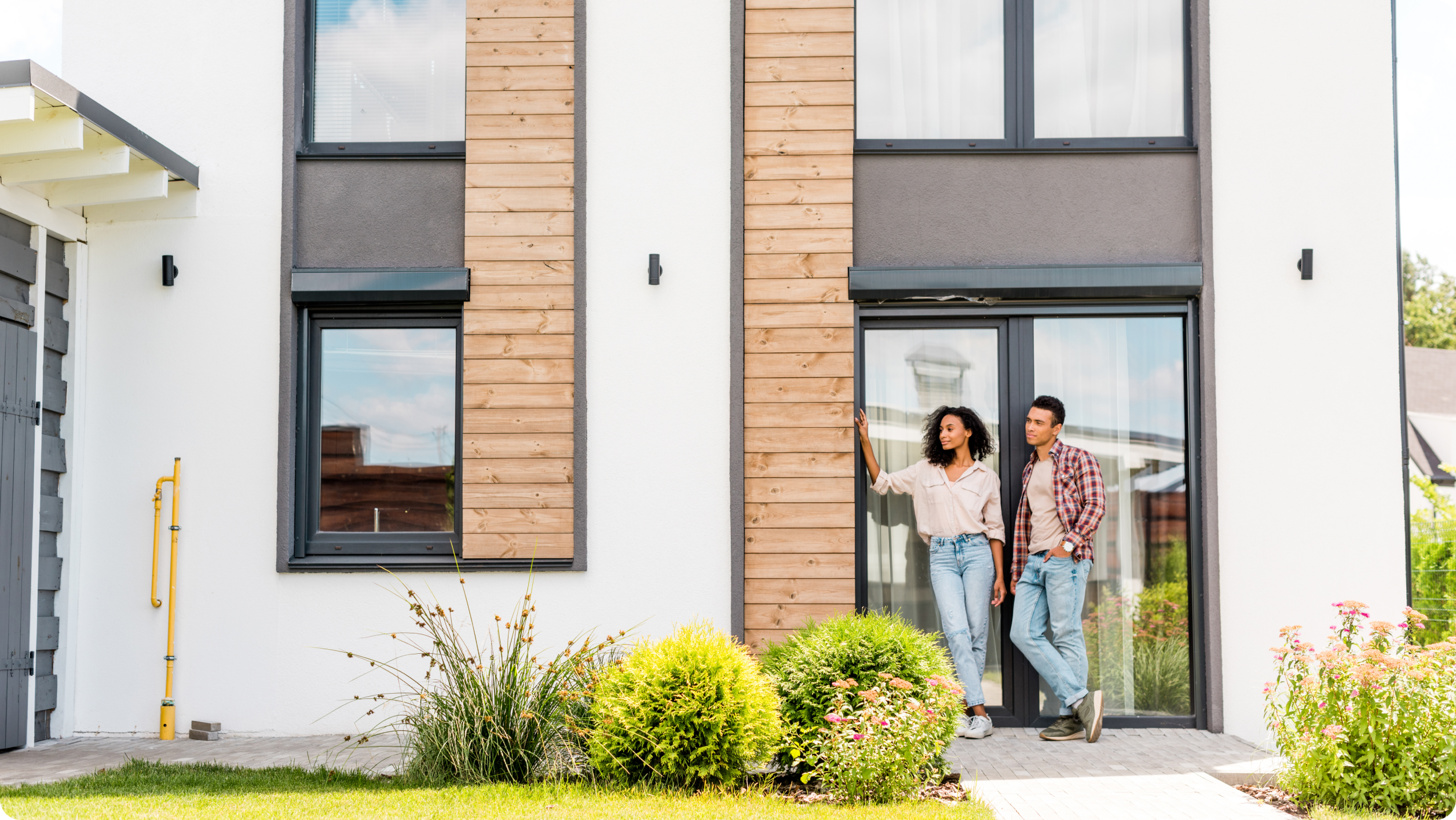 A couple stands outside a modern house with large windows and wooden panel accents, discussing how to avoid bank fees, surrounded by landscaped green bushes and plants on a sunny day.