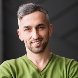 Smiling man with short gray hair and a beard, wearing a green V-neck shirt, stands against a dark, blurred background.