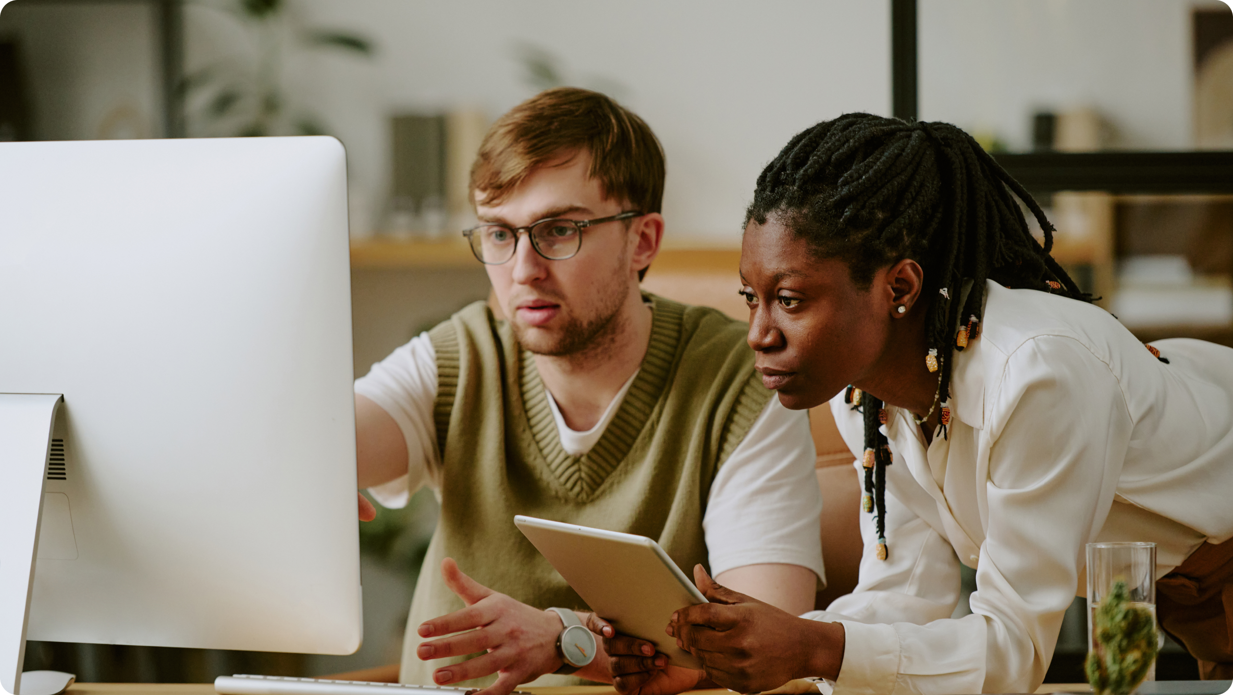 Two people are focused on a computer monitor. One is pointing at the screen, while the other holds a tablet. They appear to be in an office setting with shelves and plants in the background.