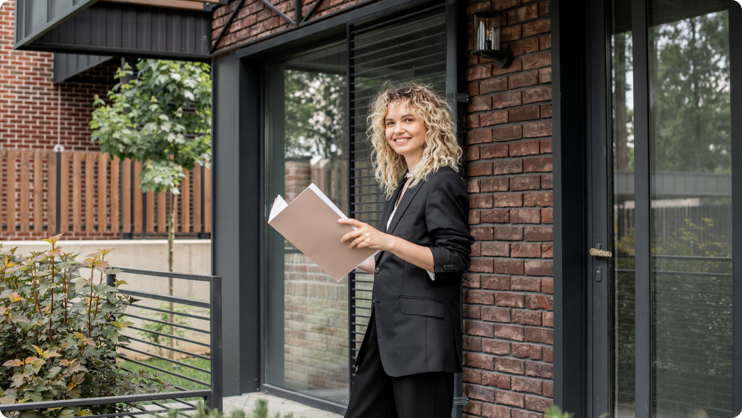 A person with curly hair is smiling and holding an open folder while standing outside a modern brick building. They are wearing a black outfit, and there is greenery in the foreground and a tree in the background.