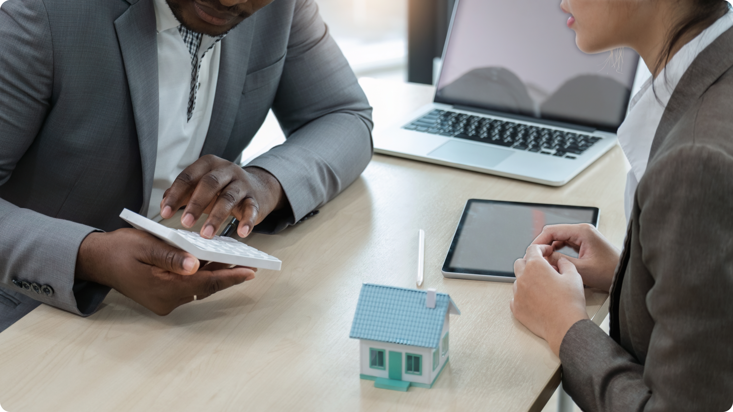 Two professionals in business attire discuss at a table with a laptop, tablet, and small house model. One uses a calculator, likely calculating security deposit interest rates, while the other listens attentively. The setting appears to be an office or conference room.