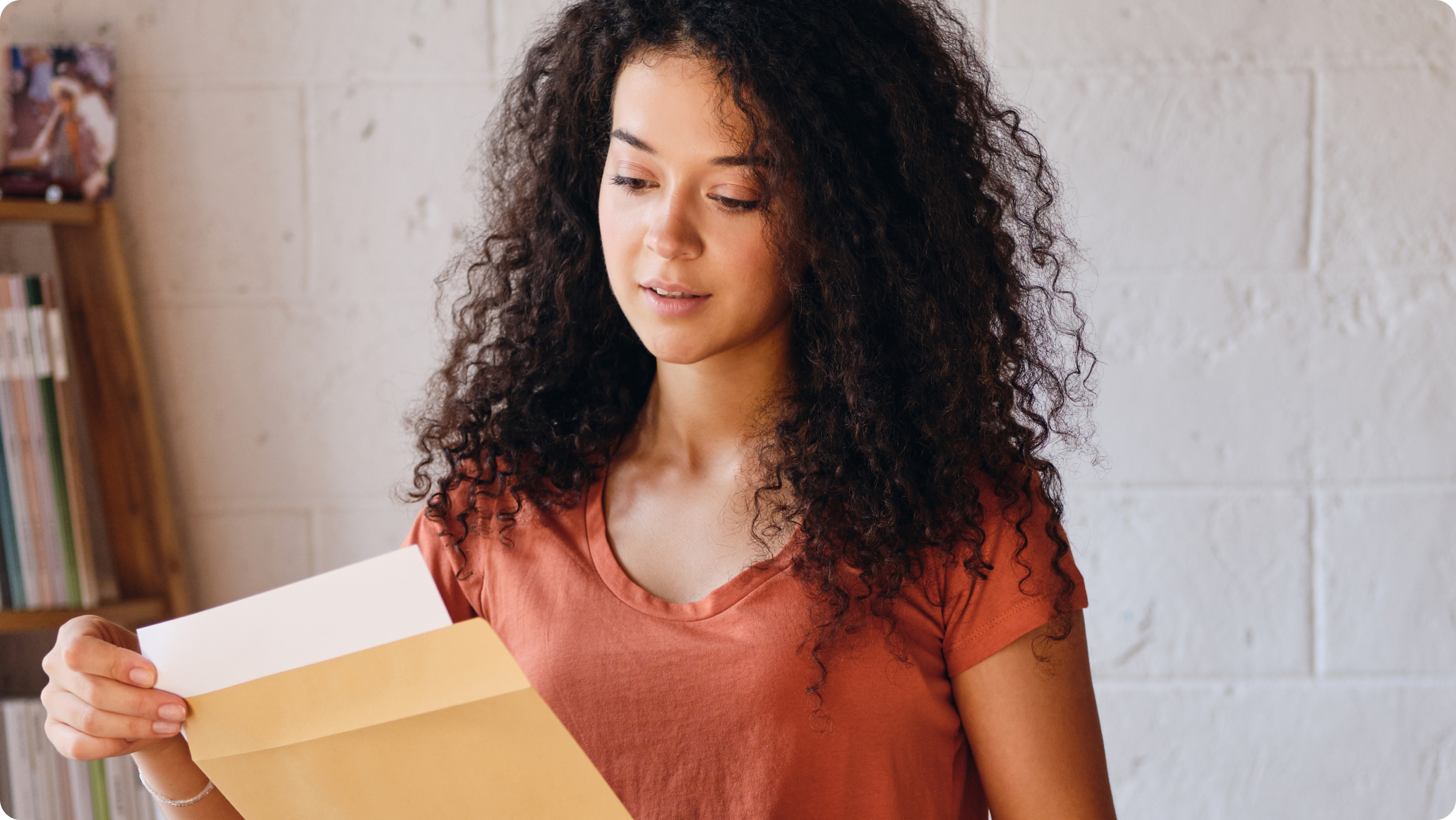 A person with long, curly hair, wearing a rust-colored shirt, stands indoors against a white brick wall. They are holding and examining an open brown envelope containing photos of rental property damage caused by a tenant's pet. Books are visible on shelves in the background.