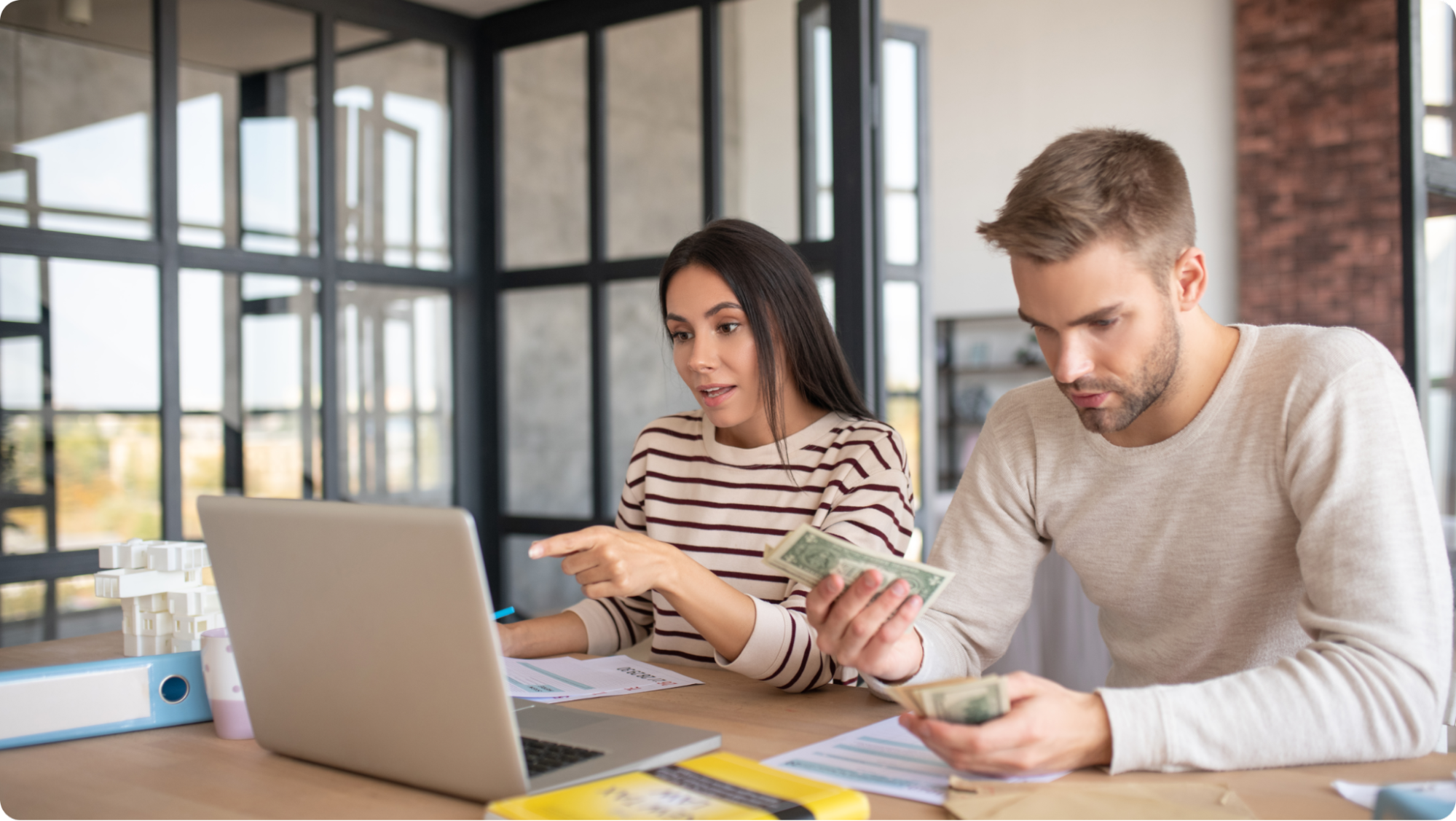 A woman and a man sit at a desk with papers, a laptop, and cash. The woman points at the laptop screen while the man counts dollar bills. They appear to be discussing finances in a modern office setting.