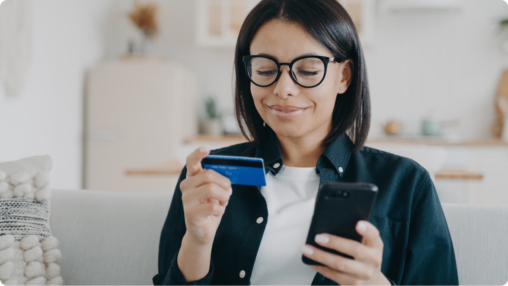 A woman wearing glasses sits on a couch, holding a credit card and a smartphone, smiling as she looks at her phone—perhaps reading about how to open an escrow account or making an online purchase.