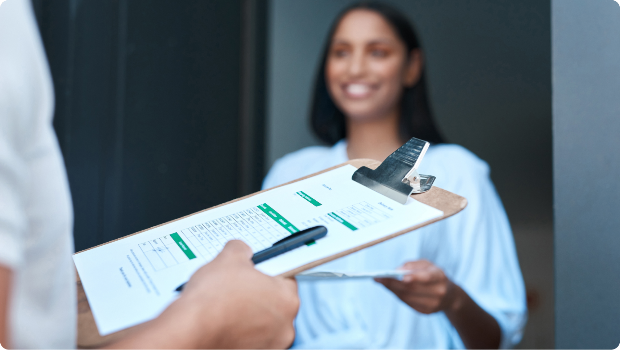 A person holding a clipboard with papers and a pen in focus. In the background, a smiling person in a light blue shirt stands near a door, holding a document. The scene suggests an inspection of rental property, ensuring no tenant pet damage has occurred.