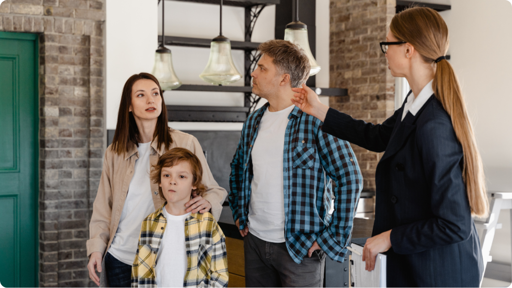 A real estate agent showing a family of three a modern home. The agent gestures towards the kitchen area. The family, including a woman, a man, and a child, listens attentively. The setting features brick walls and hanging lights.