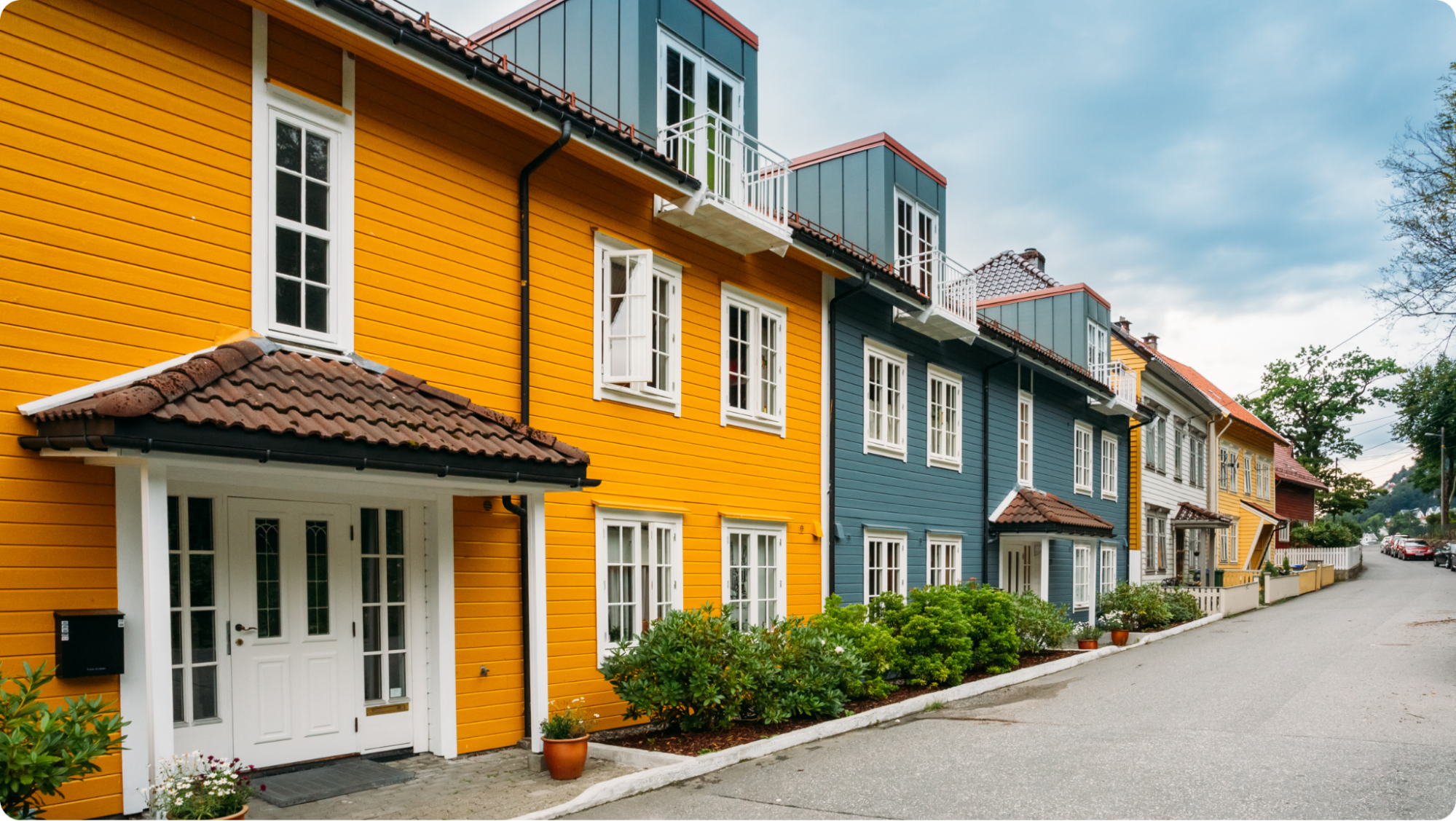 A row of colorful wooden houses with white trim and tile roofs lines a quiet street. The nearest house is bright yellow, followed by blue and beige houses, with greenery along the sidewalk under a cloudy sky.