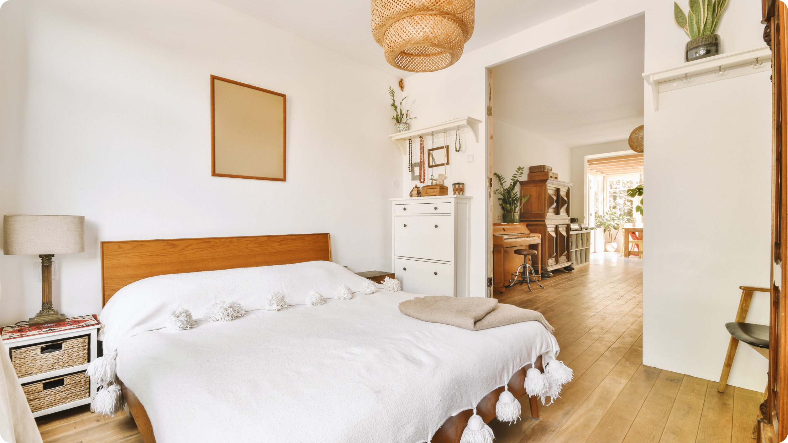 A cozy bedroom features a wooden bed with a white tassel blanket. Light-colored walls complement the small lamp on a wicker nightstand, while a white dresser stands nearby. A hallway leads to a sunlit area with a piano and plants, bringing harmony akin to knowing how to open an escrow account.