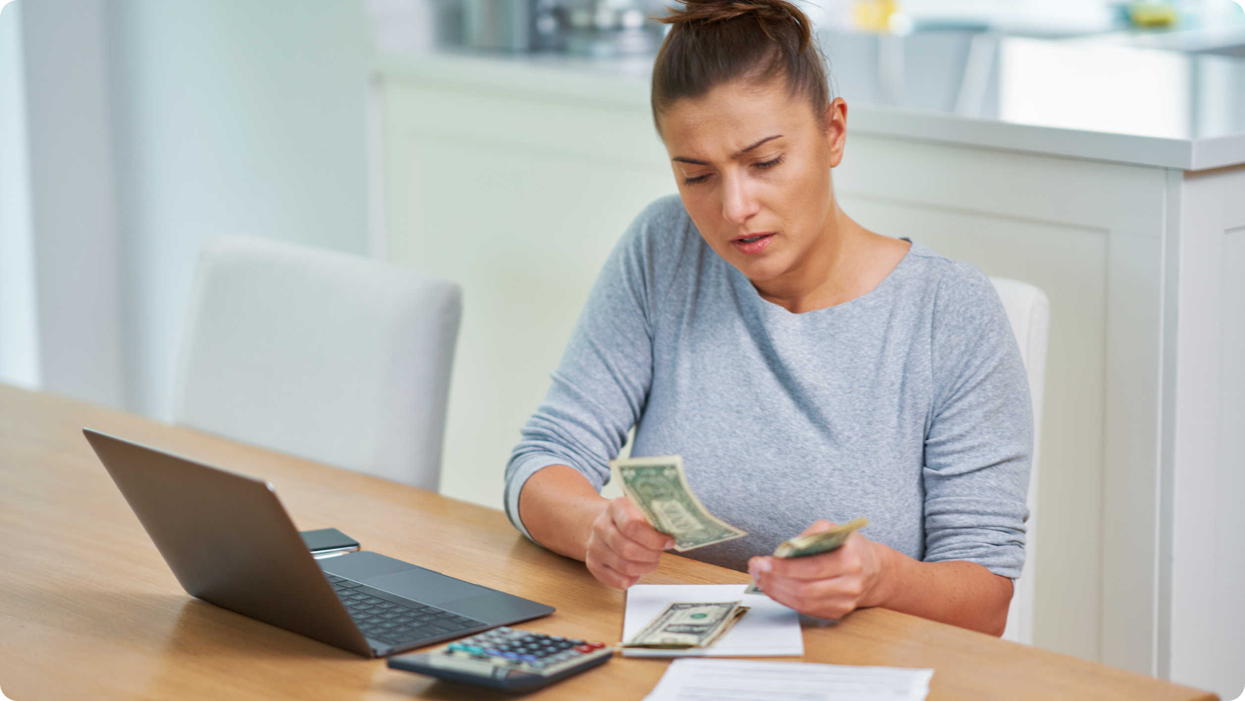 A woman sits at a table with a laptop, calculator, and notepad, counting dollar bills and looking concerned as she researches how to avoid bank fees.