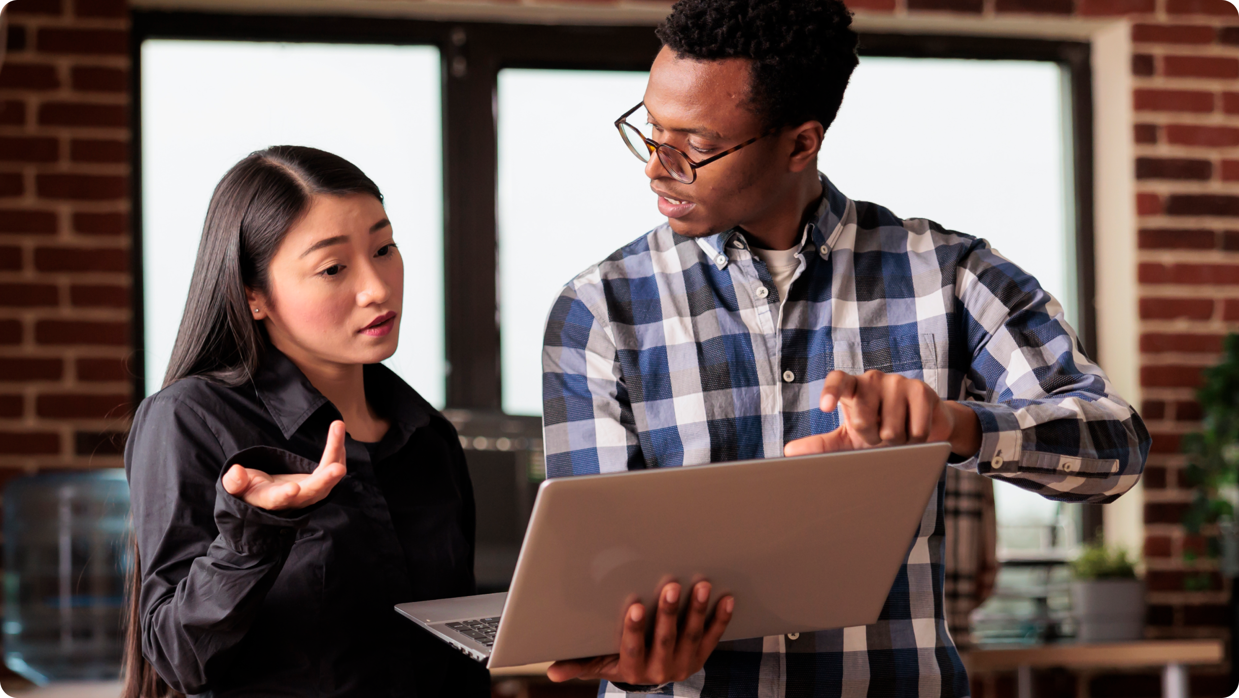 A man in a blue checkered shirt holds a laptop, gesturing at the screen. He is explaining something to a woman in a black shirt, who raises her hand in contemplation. They are standing in an office with brick walls and large windows.