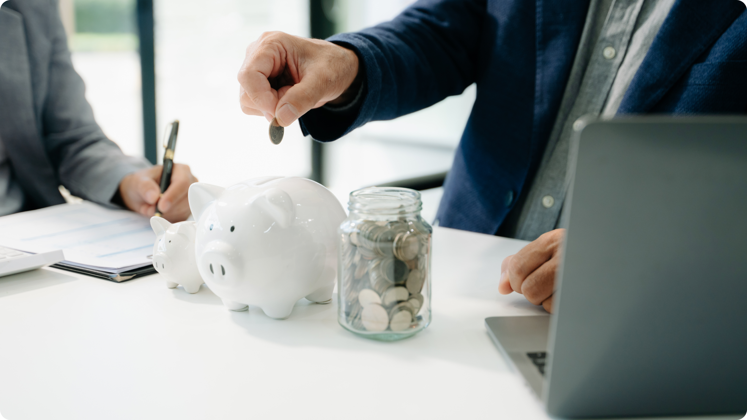 A person drops a coin into a white piggy bank on a desk, next to a jar of coins, a laptop showing rental management software, and another person writing on a clipboard nearby.