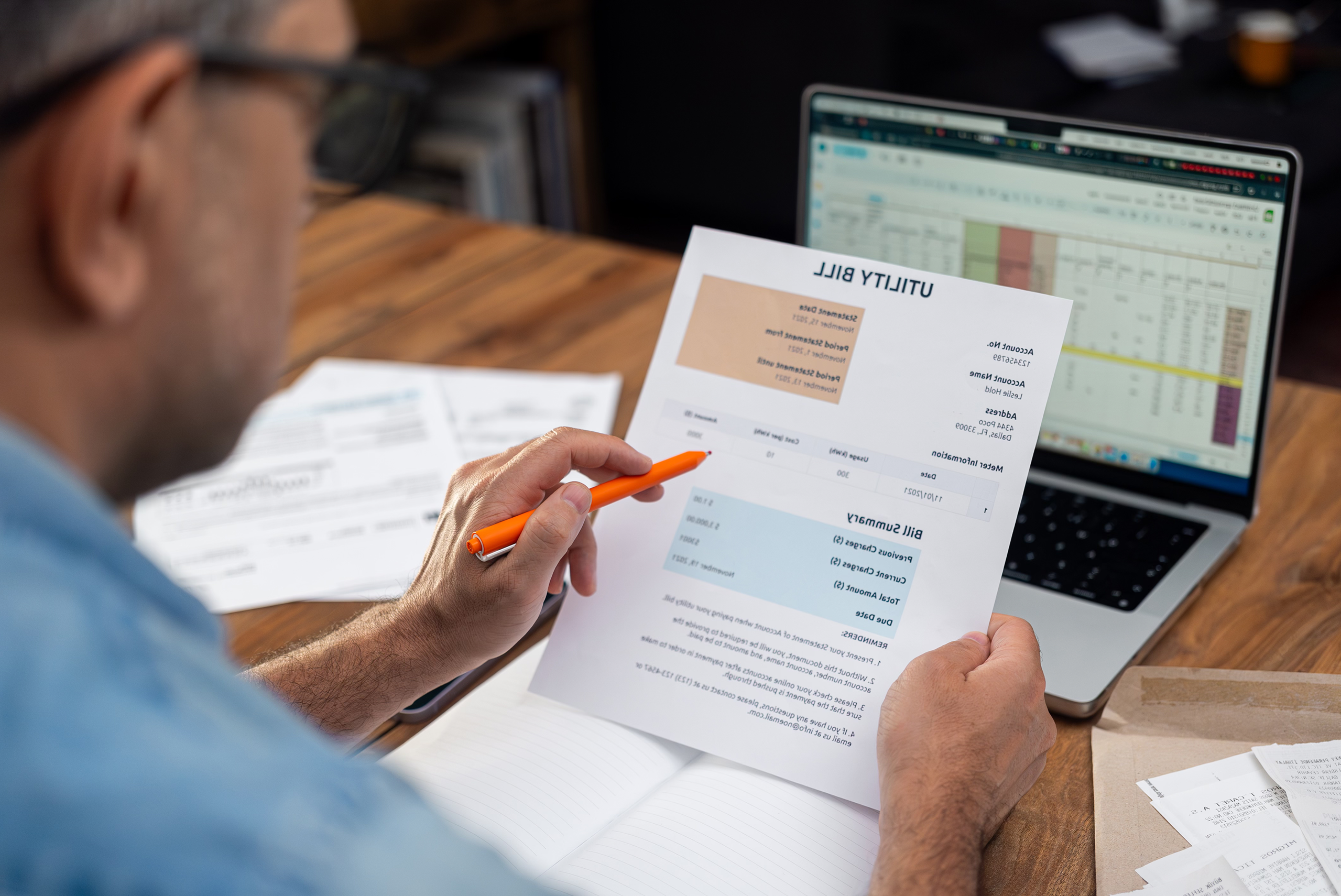 A person holding and reviewing a utility bill with a pen in hand, seated at a desk with a laptop displaying a spreadsheet and papers scattered around—considering expenses for a business vs personal bank account.