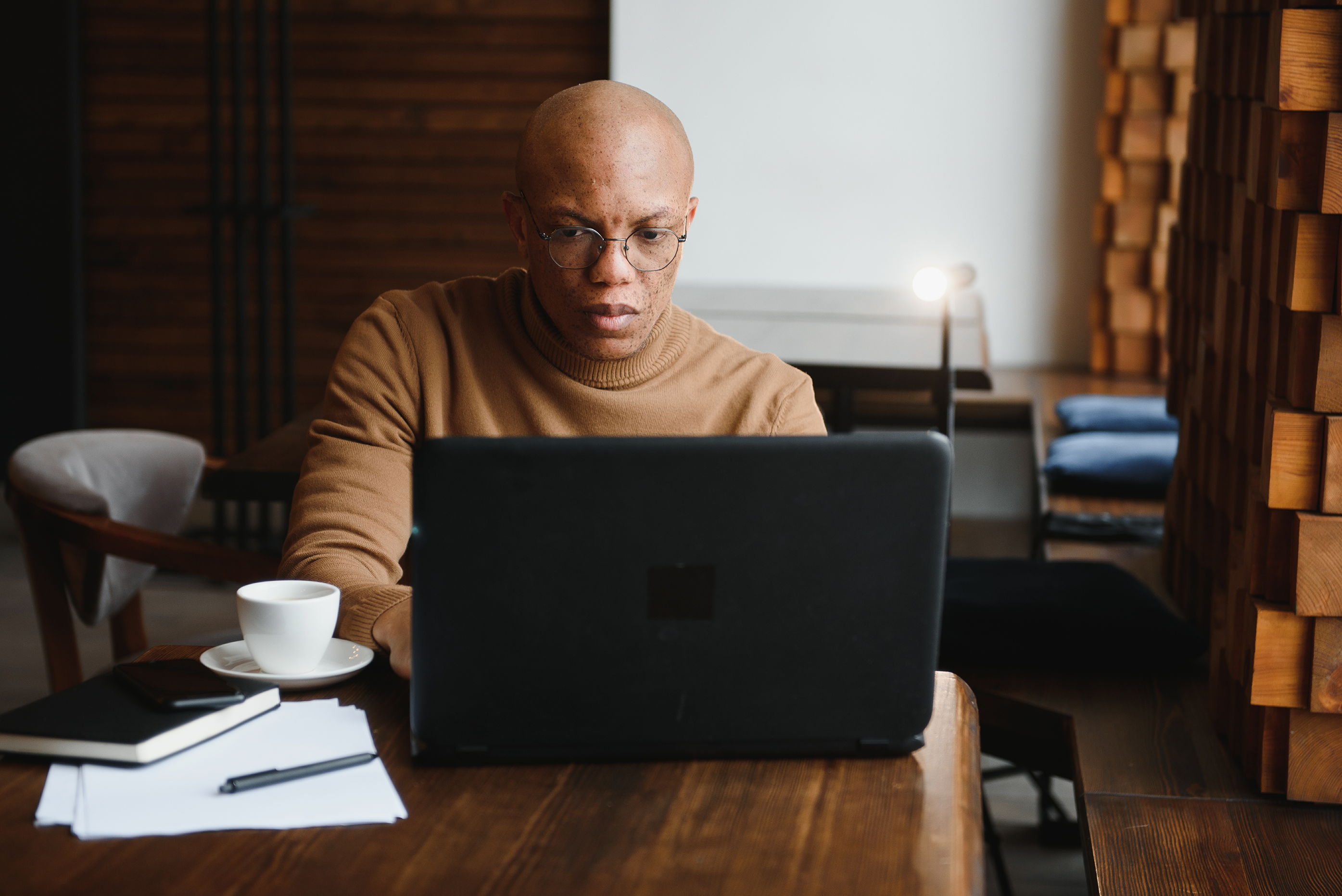 A person wearing glasses and a brown sweater sits at a wooden table, working on a laptop with rental management software. A cup of coffee, notebook, and papers with a pen are on the table in a cozy, modern setting.