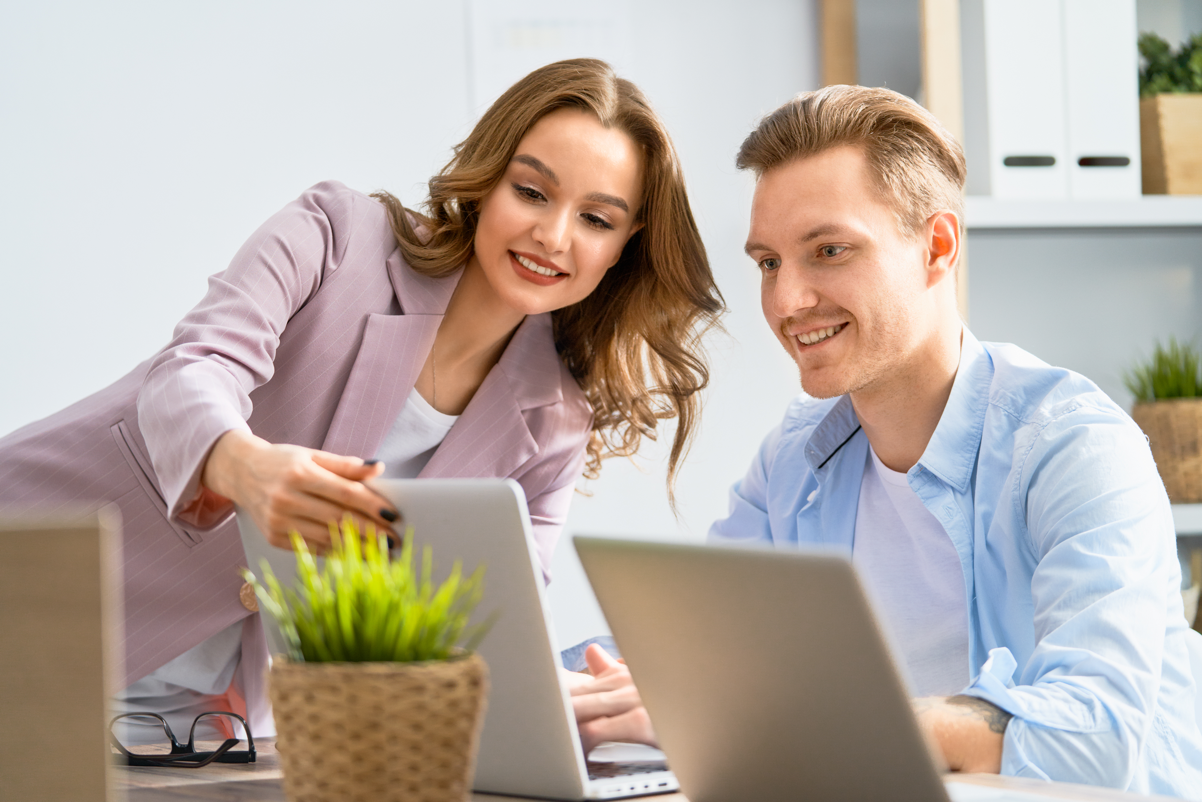 Two colleagues smiling and looking at a laptop screen together in a bright office setting, discussing topics like business vs personal bank account​. The woman points at the screen as the man listens, both appearing engaged and collaborative. A plant sits on the desk.