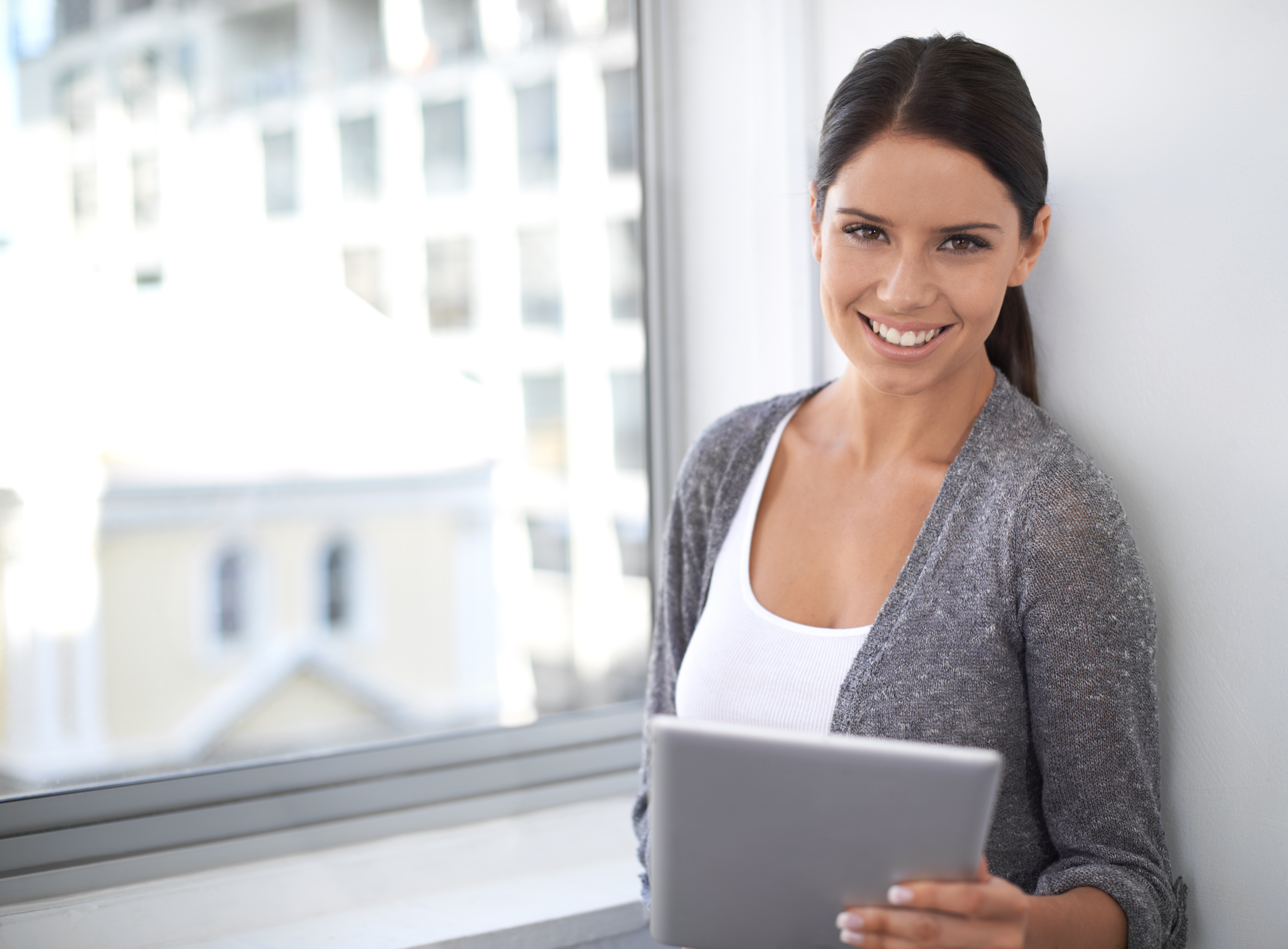 A woman with dark hair, wearing a gray cardigan and white top, smiles while holding a tablet near a window. She researches business vs personal bank account​ options against the backdrop of a blurred cityscape.