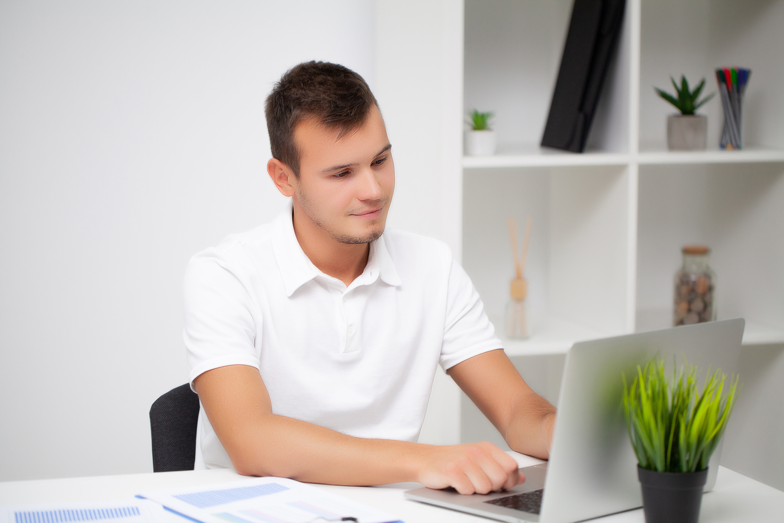A young man in a white polo shirt sits at a desk, working on his laptop with rental management software. Papers, a green plant, and office supplies are on the desk, with shelves and potted plants in the background.