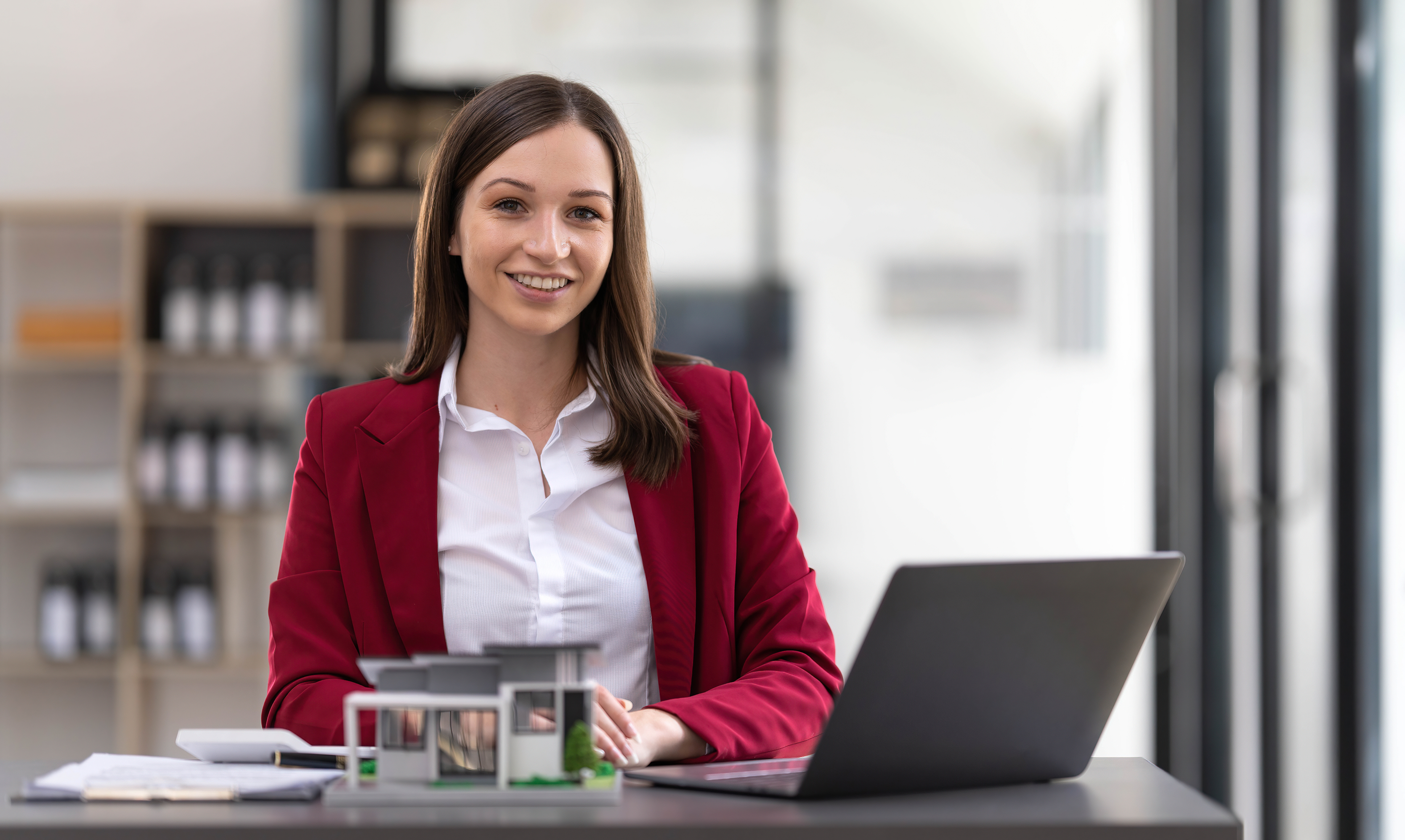 A smiling woman in a red blazer sits at a desk with a laptop, paperwork, and a modern house model, perhaps reviewing business vs personal bank account details for her real estate work. Shelves and bright windows are visible in the background.