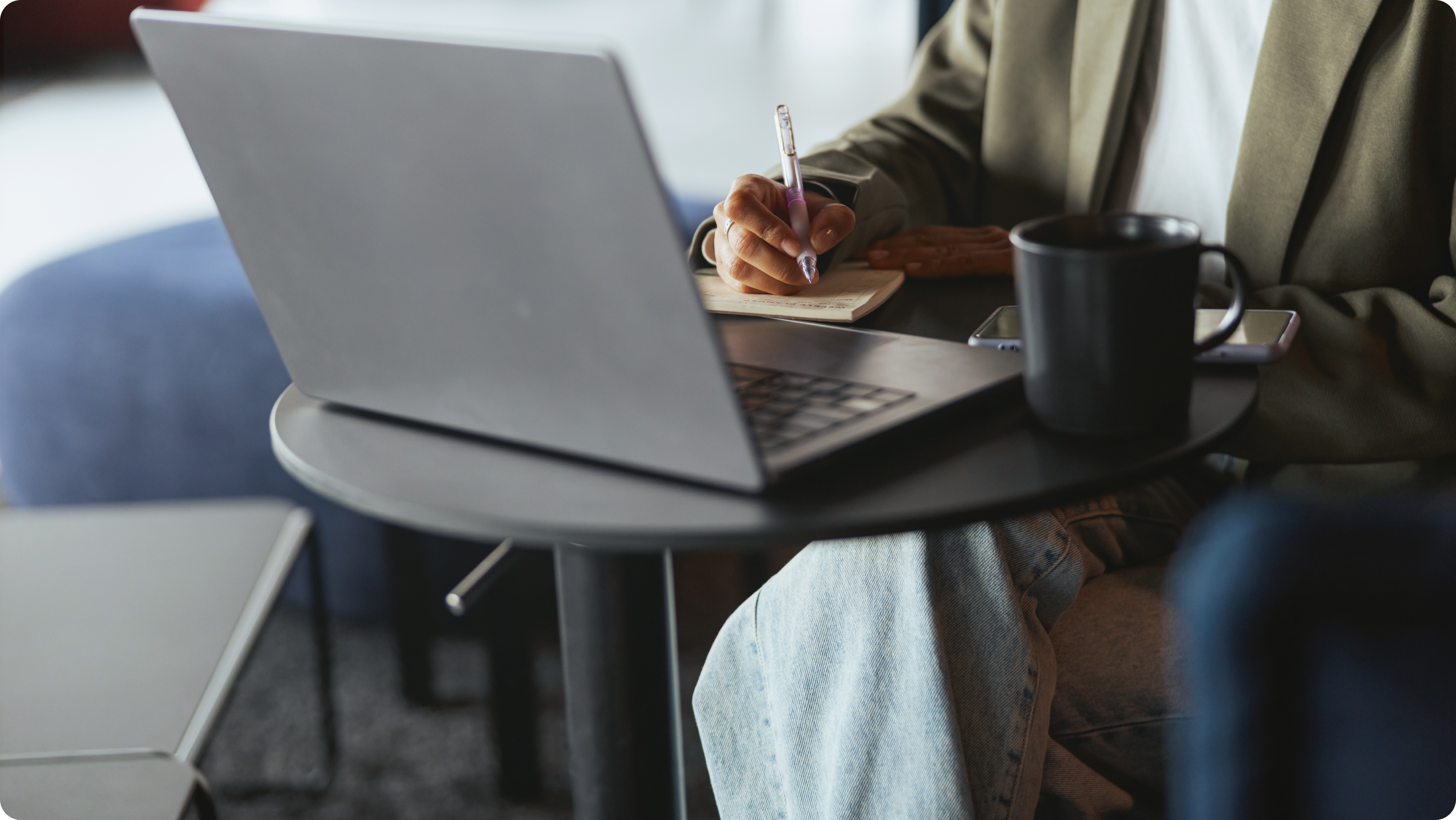 A person sits at a round table with a laptop, writing in a notebook about business vs personal bank account​ considerations. A black mug and a smartphone rest on the table in this casual, modern setting.