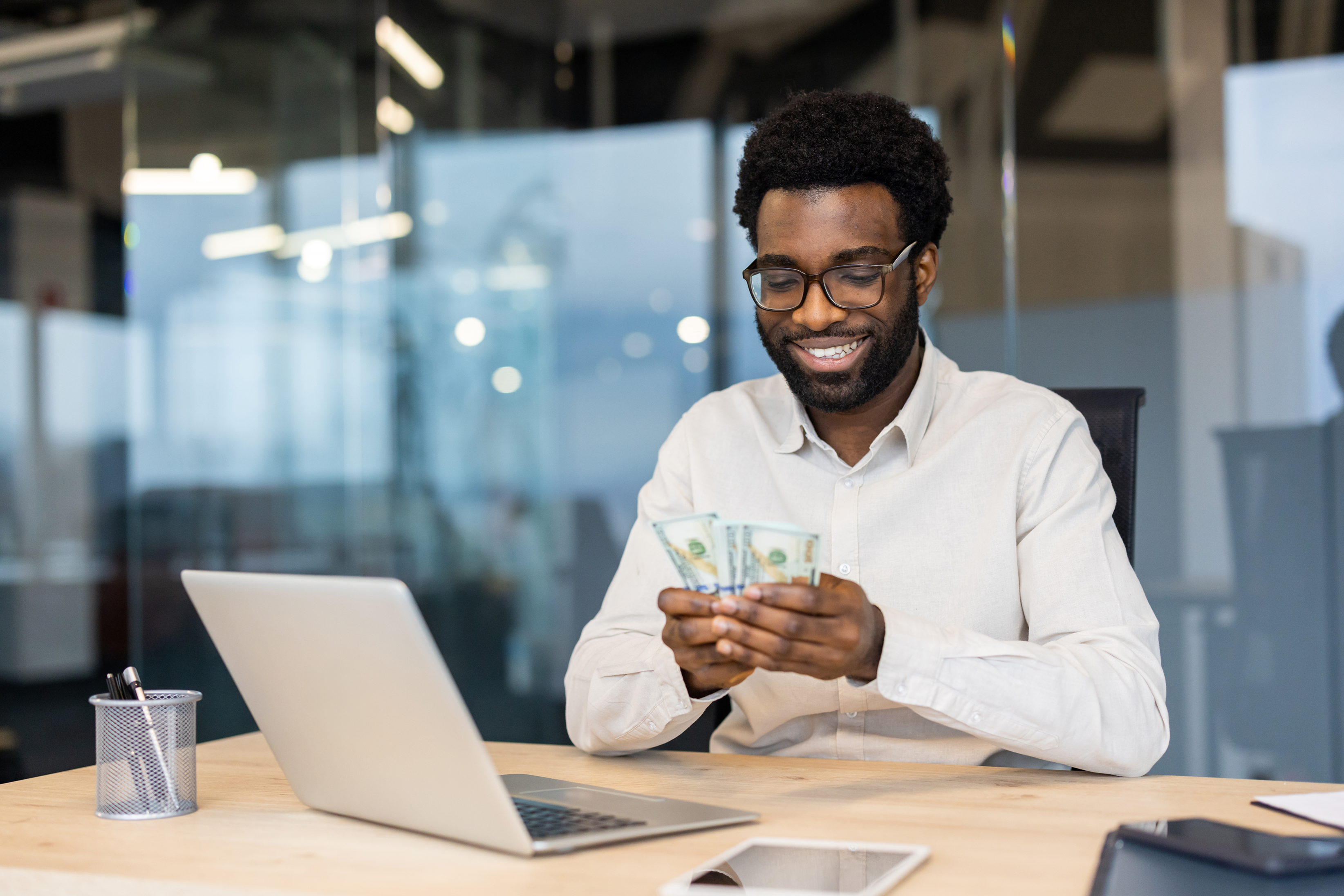 A man wearing glasses and a white shirt sits at a desk with a laptop, smiling as he counts dollar bills—perhaps weighing the benefits of a business vs personal bank account. Office supplies and a tablet sit on the desk in this modern office setting.