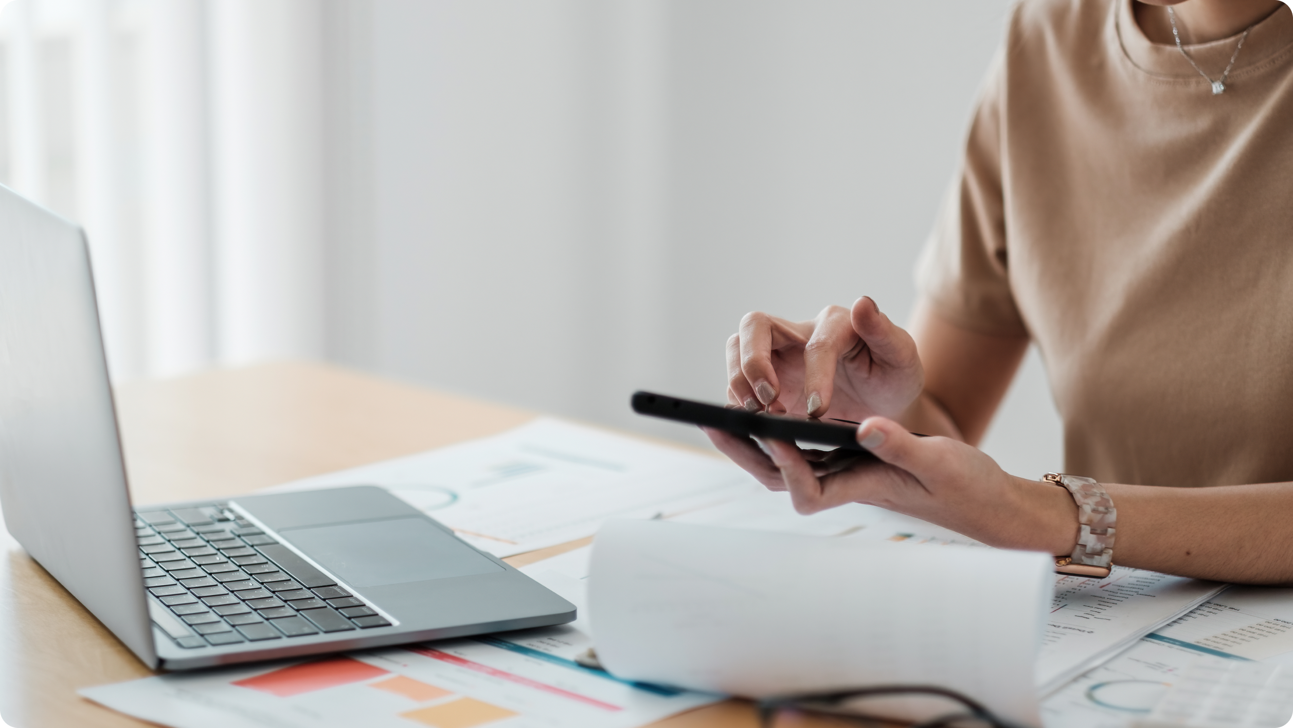 A person wearing a brown shirt uses a calculator at a desk with an open laptop, financial documents, and charts—possibly analyzing the differences between a business vs personal bank account for accounting or data analysis purposes.