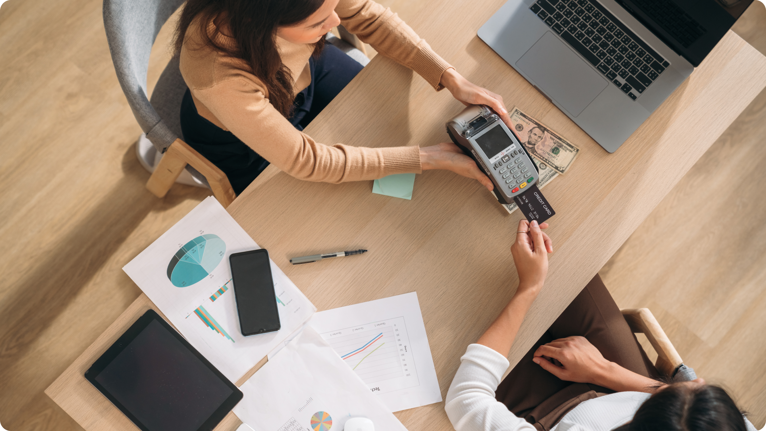 Two people sit at a desk with a laptop, tablet, papers, and cash. One hands a card to the other using a payment terminal, suggesting a business or financial transaction—possibly streamlined by rental management software.