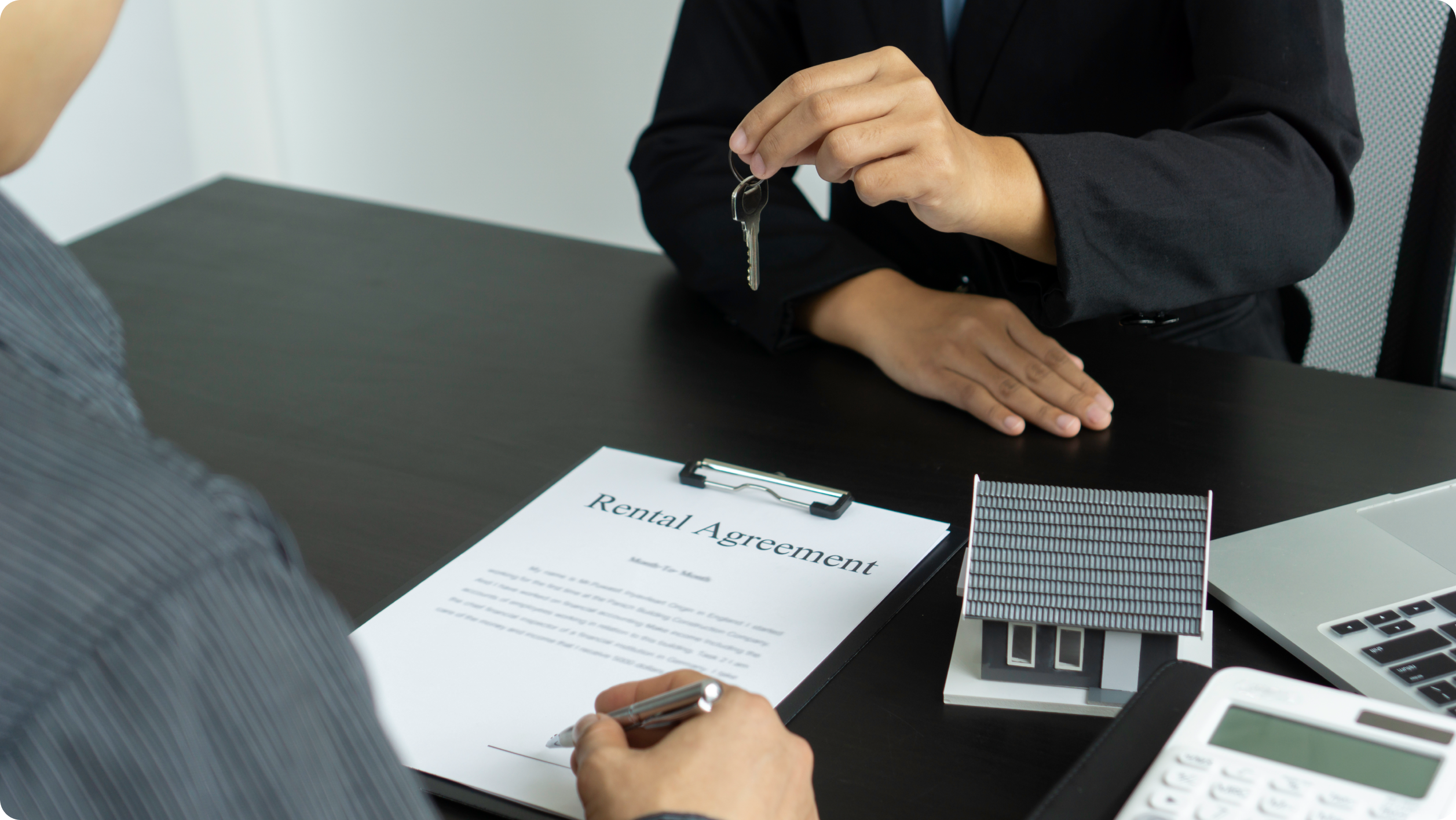 A person hands over a key to another signing a rental agreement on a clipboard, with a small model house and calculator on the table—highlighting important details like choosing between a business vs personal bank account​.