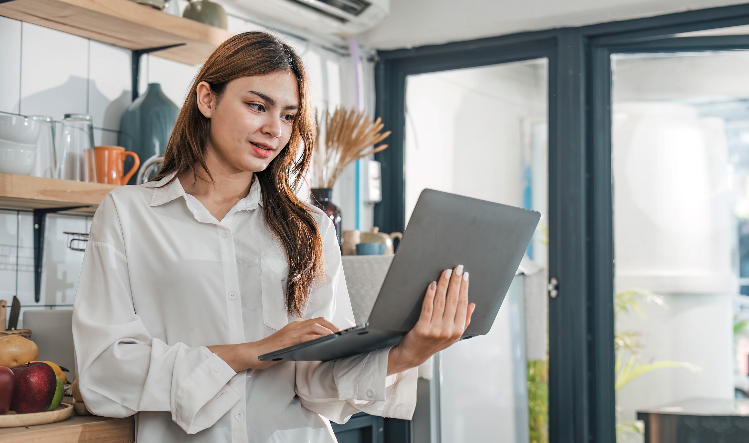 A woman in a white shirt stands in a kitchen, holding and looking at a laptop—perhaps researching business vs personal bank account options. Shelves with dishes, glassware, and fruit are visible behind her, lit by large windows.