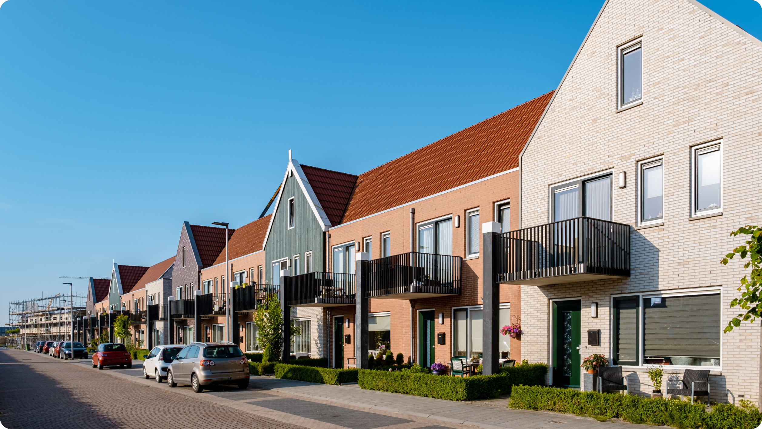 A row of modern, colorful townhouses with pitched roofs and balconies lines the street, where parked cars suggest residents balancing business vs personal bank account needs under a clear blue sky on a sunny day.