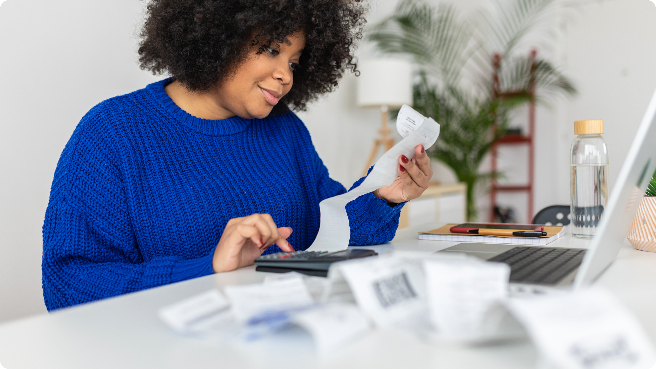 A woman in a blue sweater sits at a desk with receipts, using a calculator and looking at her laptop, appearing to manage her finances—perhaps deciding between a business vs personal bank account. Plants and office supplies are visible in the background.