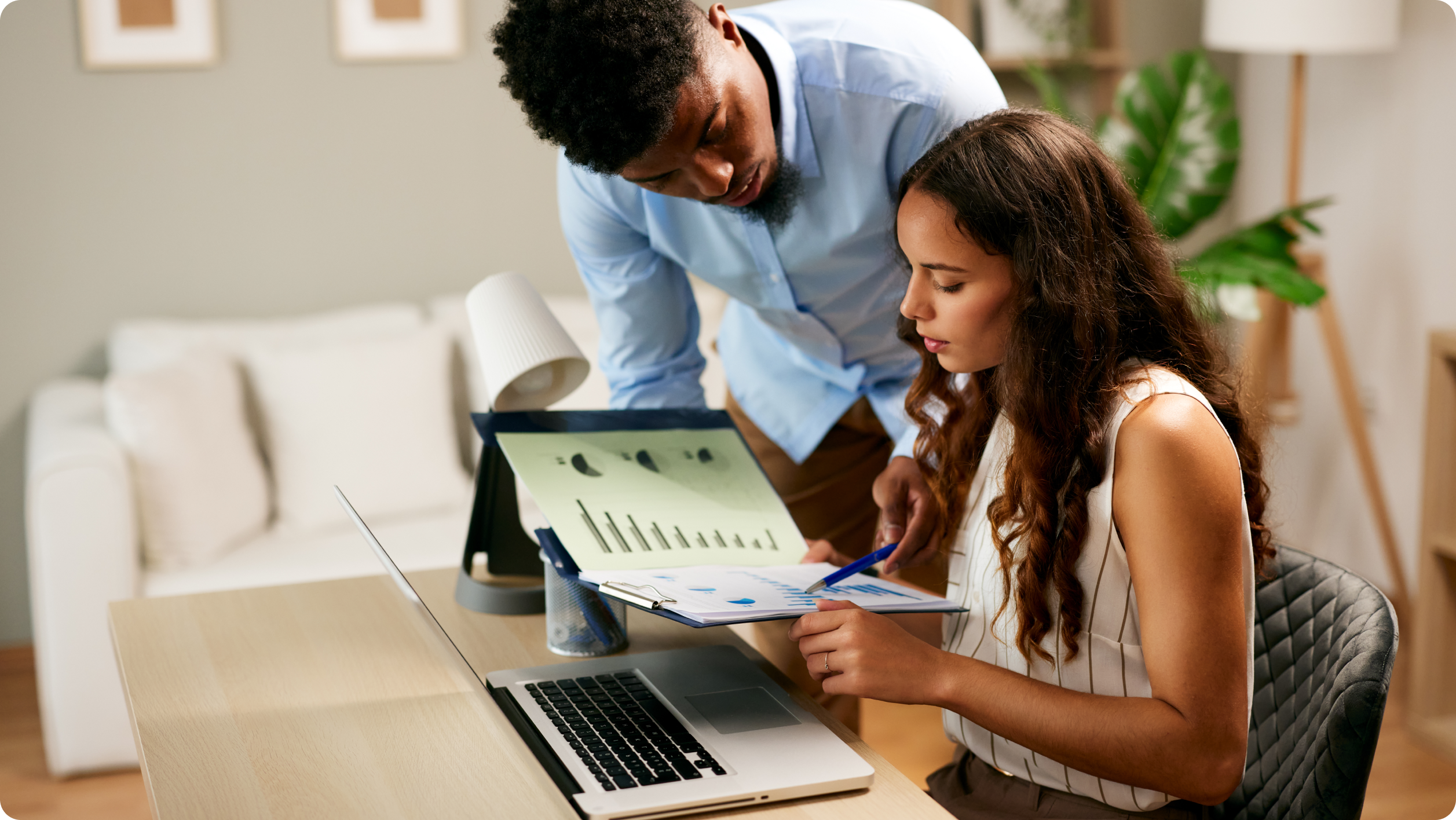 A woman sits at a desk with a laptop, holding papers with graphs, while a man stands beside her, leaning over and discussing the documents. They appear to be collaborating on rental management software in an office setting.
