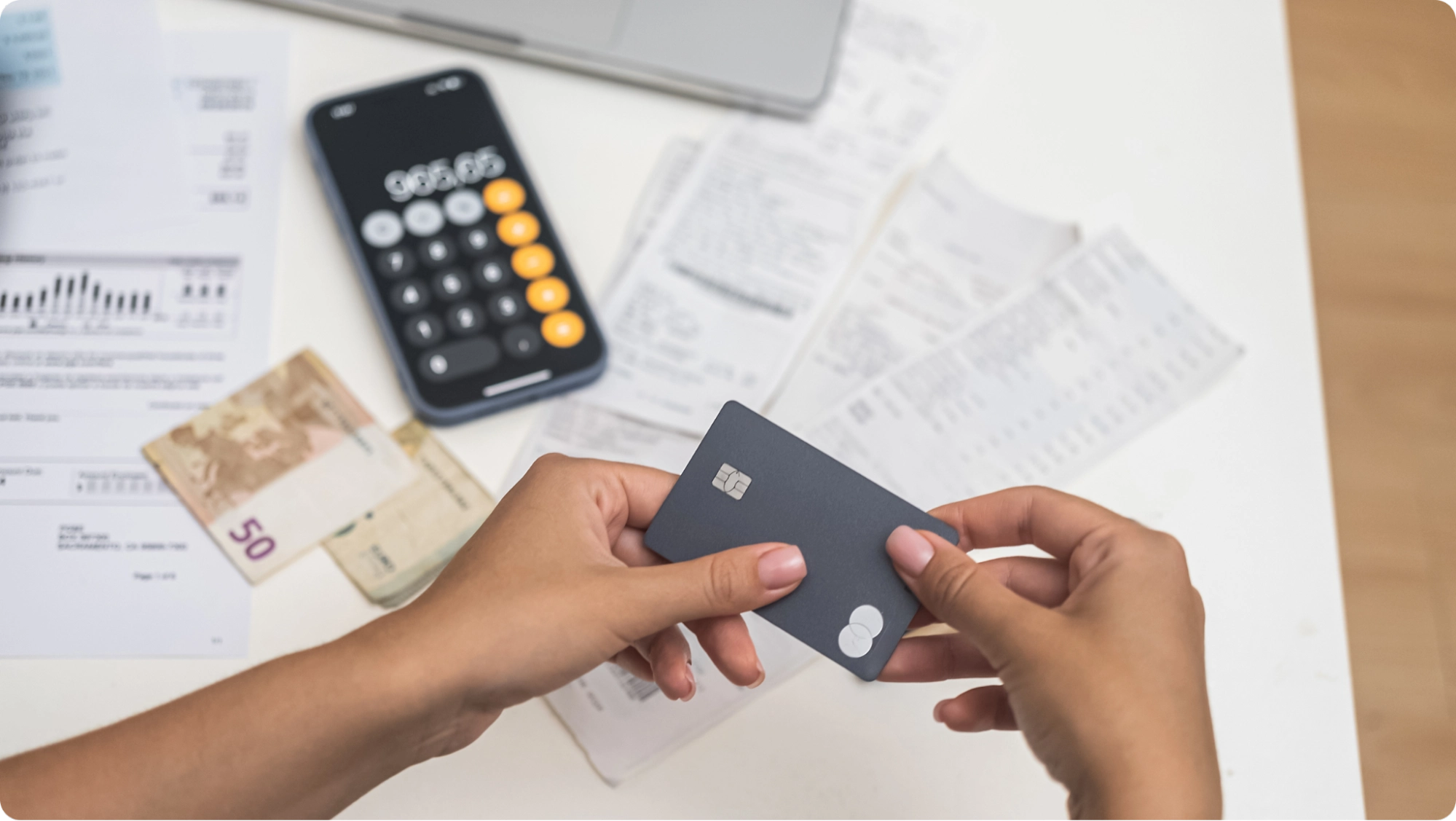 A person holds a credit card over a table with receipts, a calculator app on a smartphone, euro bills, and financial documents, suggesting budgeting or tracking expenses like security deposit rent.