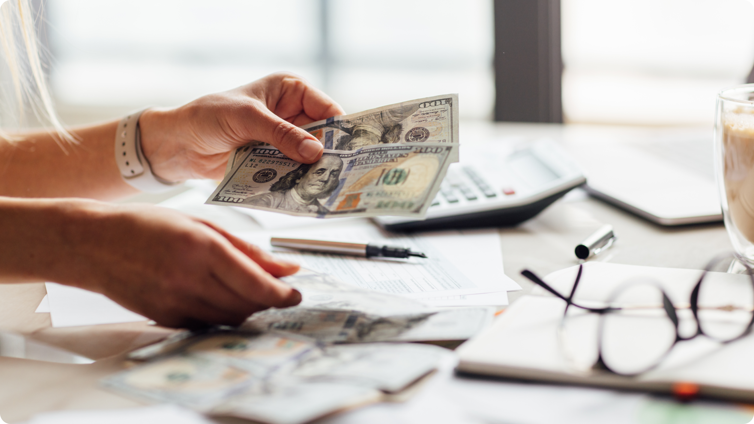 A person counts $100 bills at a desk with documents, a pen, calculator, eyeglasses, and coffee—perhaps reviewing their finances or exploring no fee checking accounts​.
