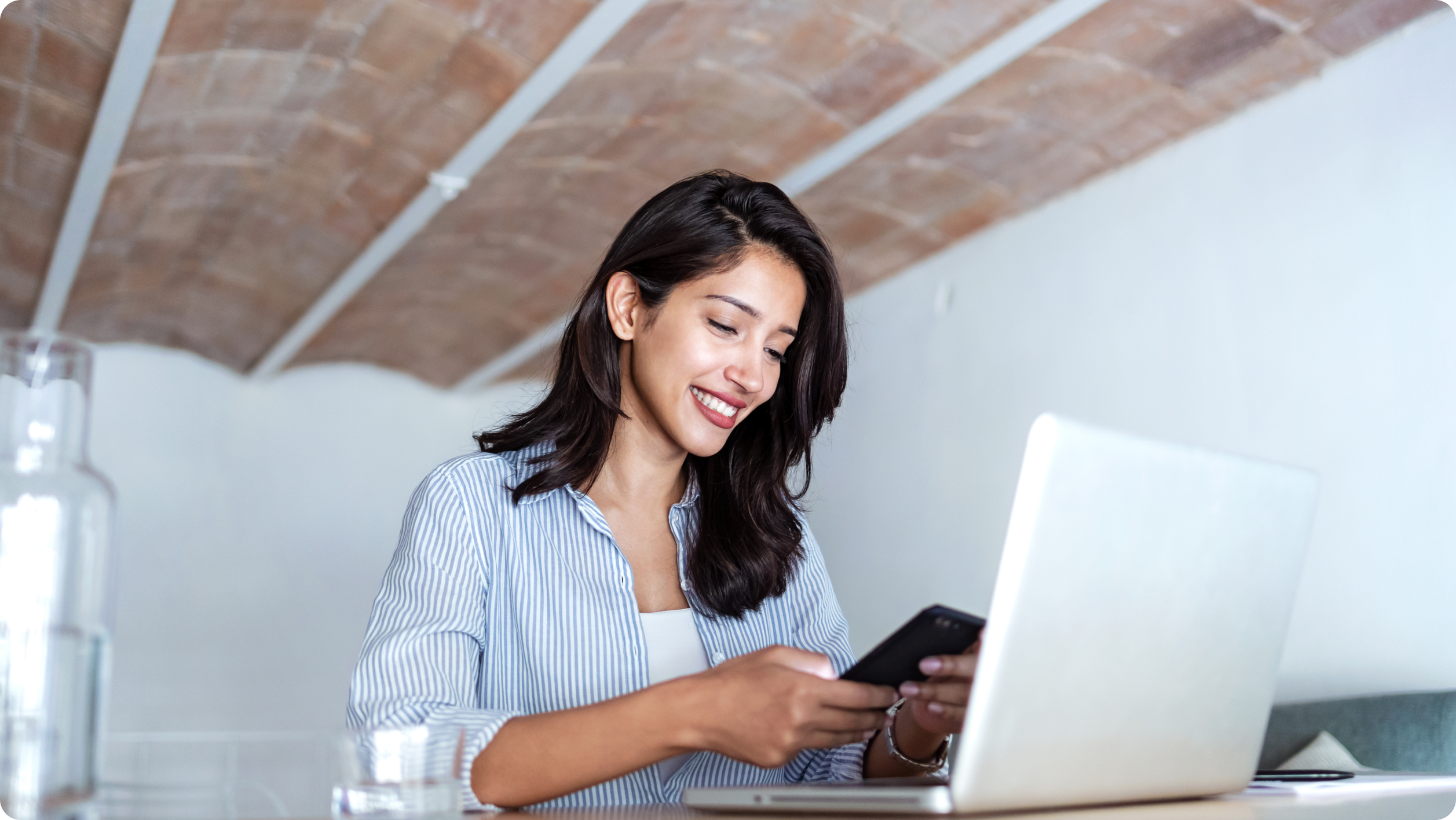 A woman sits at a table with a laptop, smiling while looking at her smartphone. She wears a blue striped shirt and there is a glass of water and a glass pitcher nearby. The setting appears to be bright and modern.