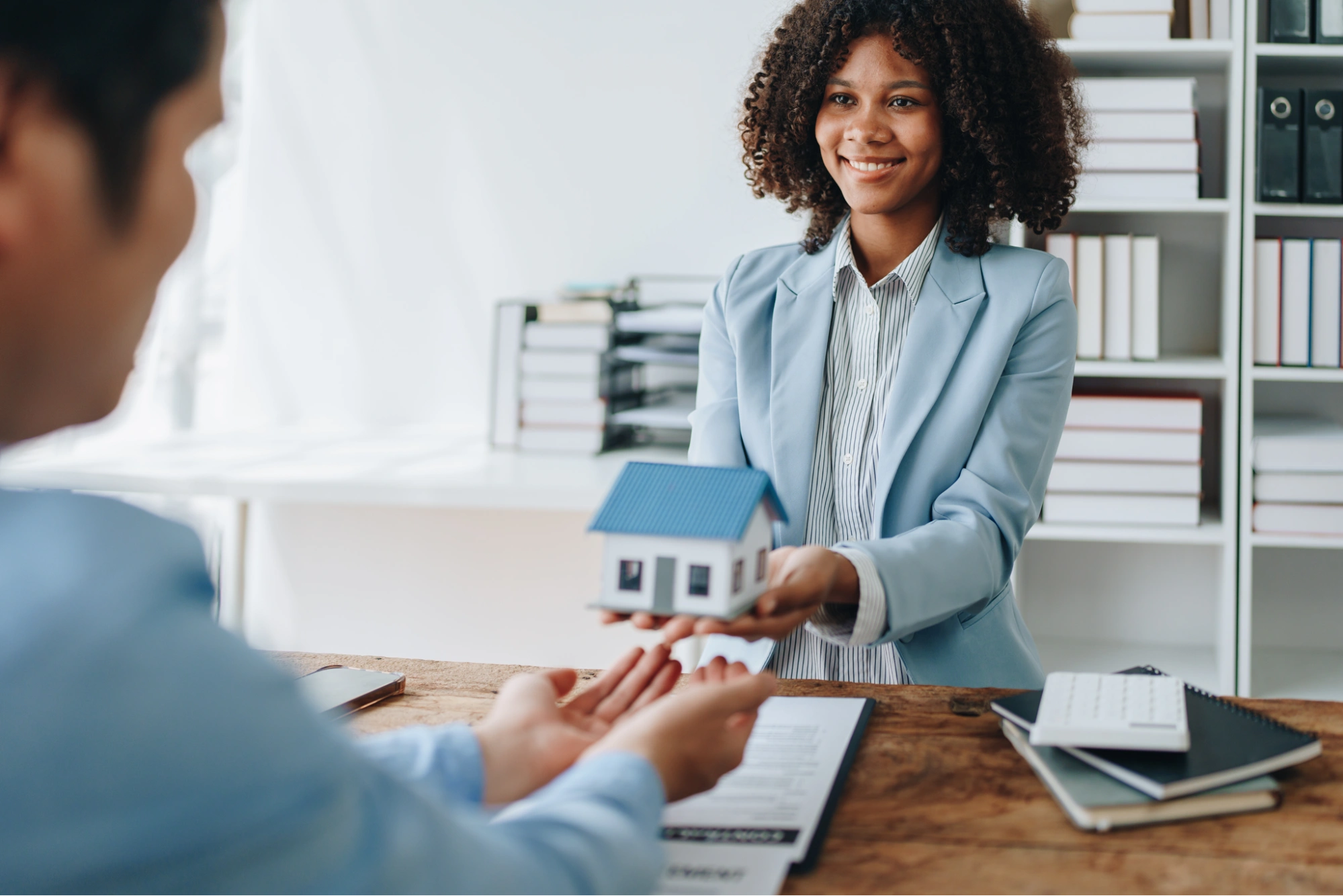 A smiling woman in a blue blazer hands a small model house to another person across a desk, suggesting a real estate or home-buying transaction—perhaps highlighting the importance of using a business vs personal bank account​ in such deals.