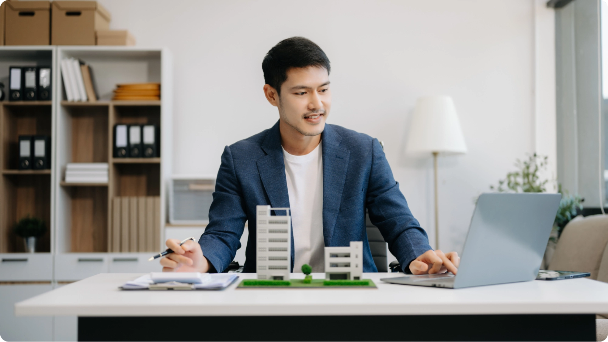 A man in a blue blazer sits at a desk with architectural models, working on a laptop and holding a pen, perhaps reviewing documents about business vs personal bank account​ options. Shelves, a lamp, and office supplies are visible in the background.